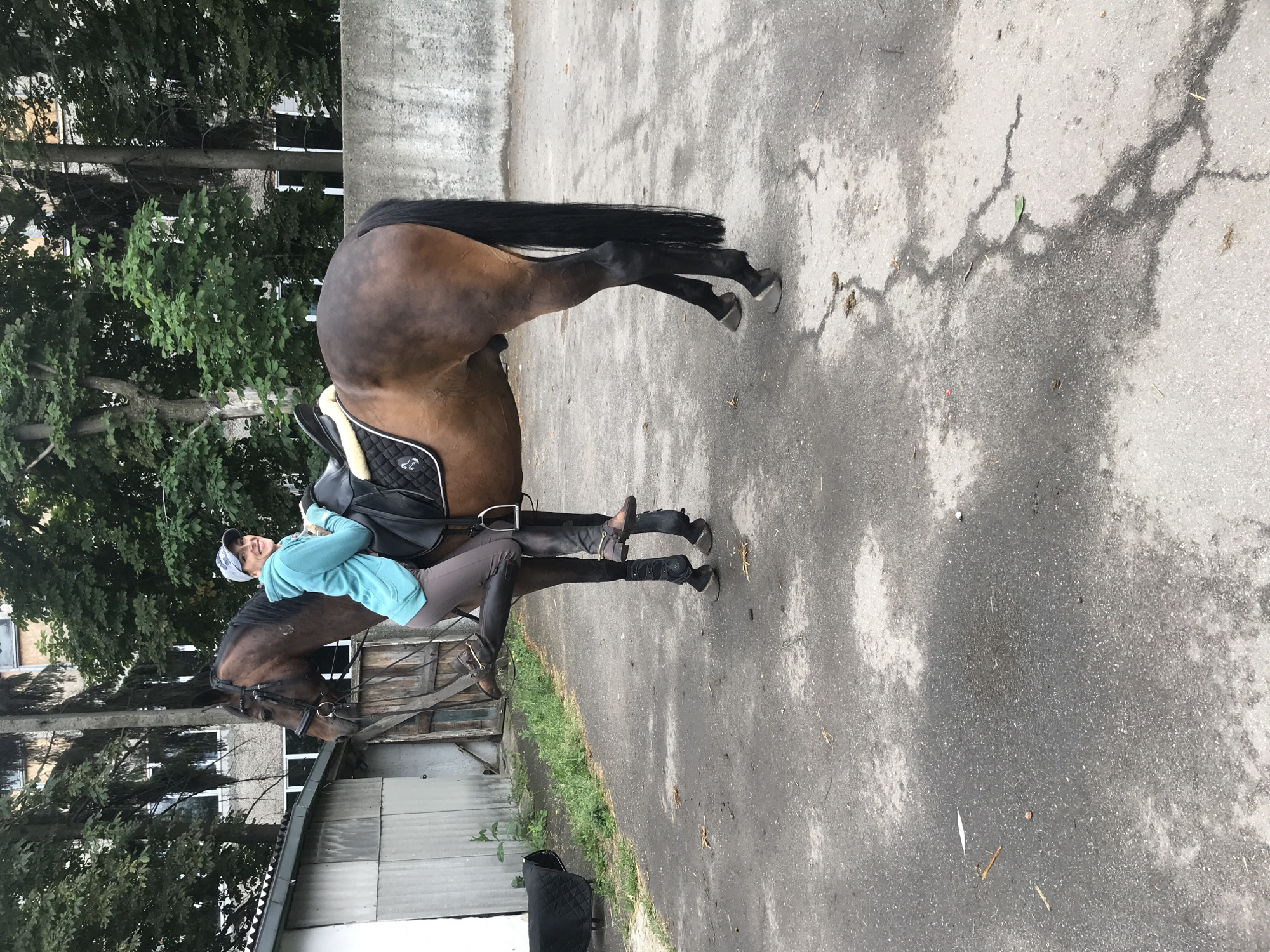 The image shows a woman riding a brown horse. The woman is wearing a light blue long-sleeved shirt, dark riding pants, and brown boots. She also has a baseball cap on her head and is smiling at the camera. The horse is equipped with a saddle and bridle. They are on an asphalt surface, possibly a road or a stable yard, with trees and a building in the background. The scene appears to be outdoors, and the lighting suggests it is daytime, likely overcast or late afternoon given the diffused light. The overall mood is casual and happy. The location context is Kharkiv, Ukraine. There is no visible text in the image.