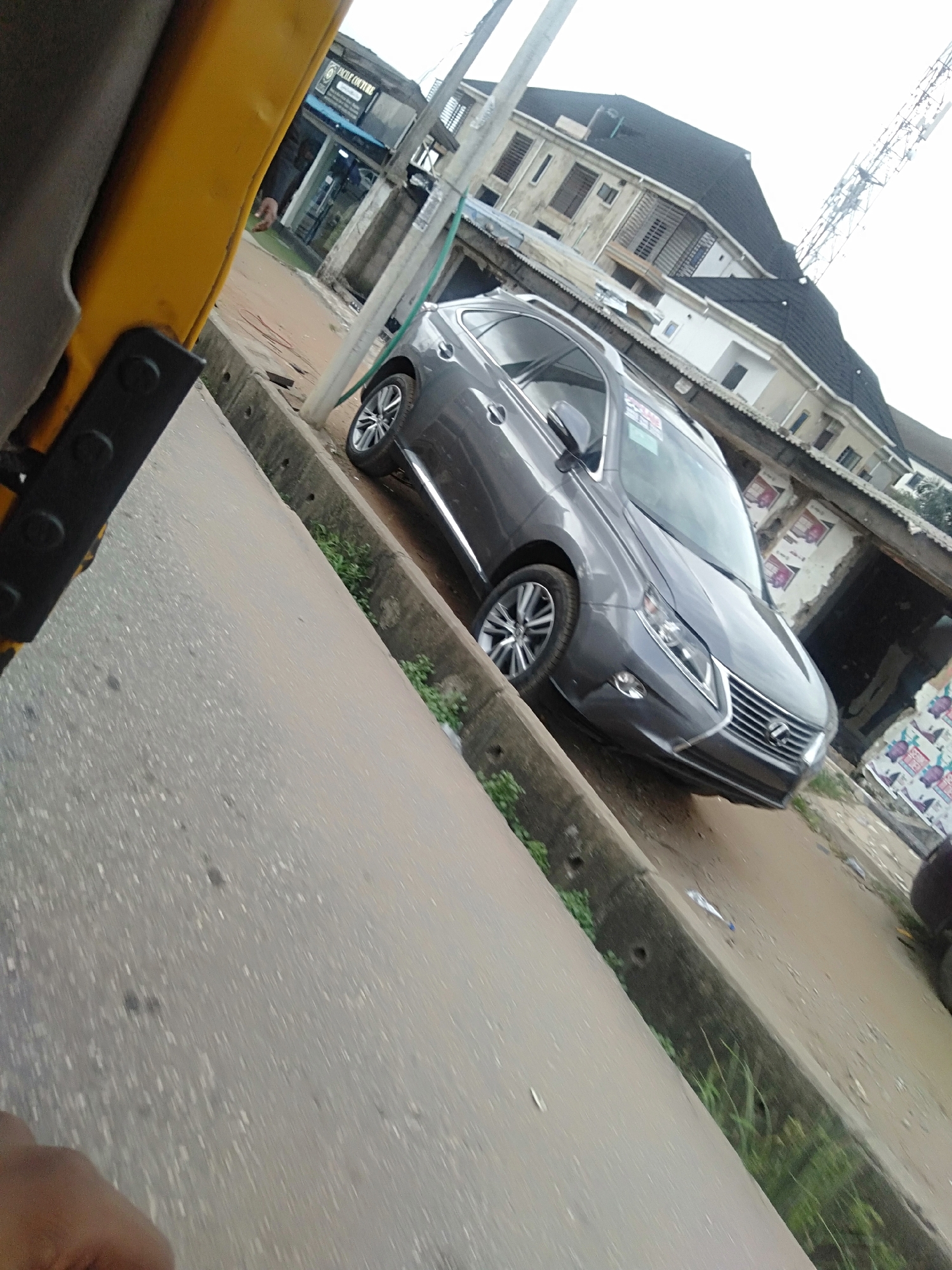 The image shows a gray Lexus RX parked on a dirt shoulder next to a road in Shangisha, Nigeria. The car is facing forward and has a "For Sale" sign in the window. The front of a yellow vehicle is visible on the left side of the image. The road is made of gray concrete. There is a gutter filled with some grass between the car and road. In the background, there are several buildings with signs and advertisements. A cell tower can be seen in the distance. The photo appears to have been taken from the inside of another vehicle.