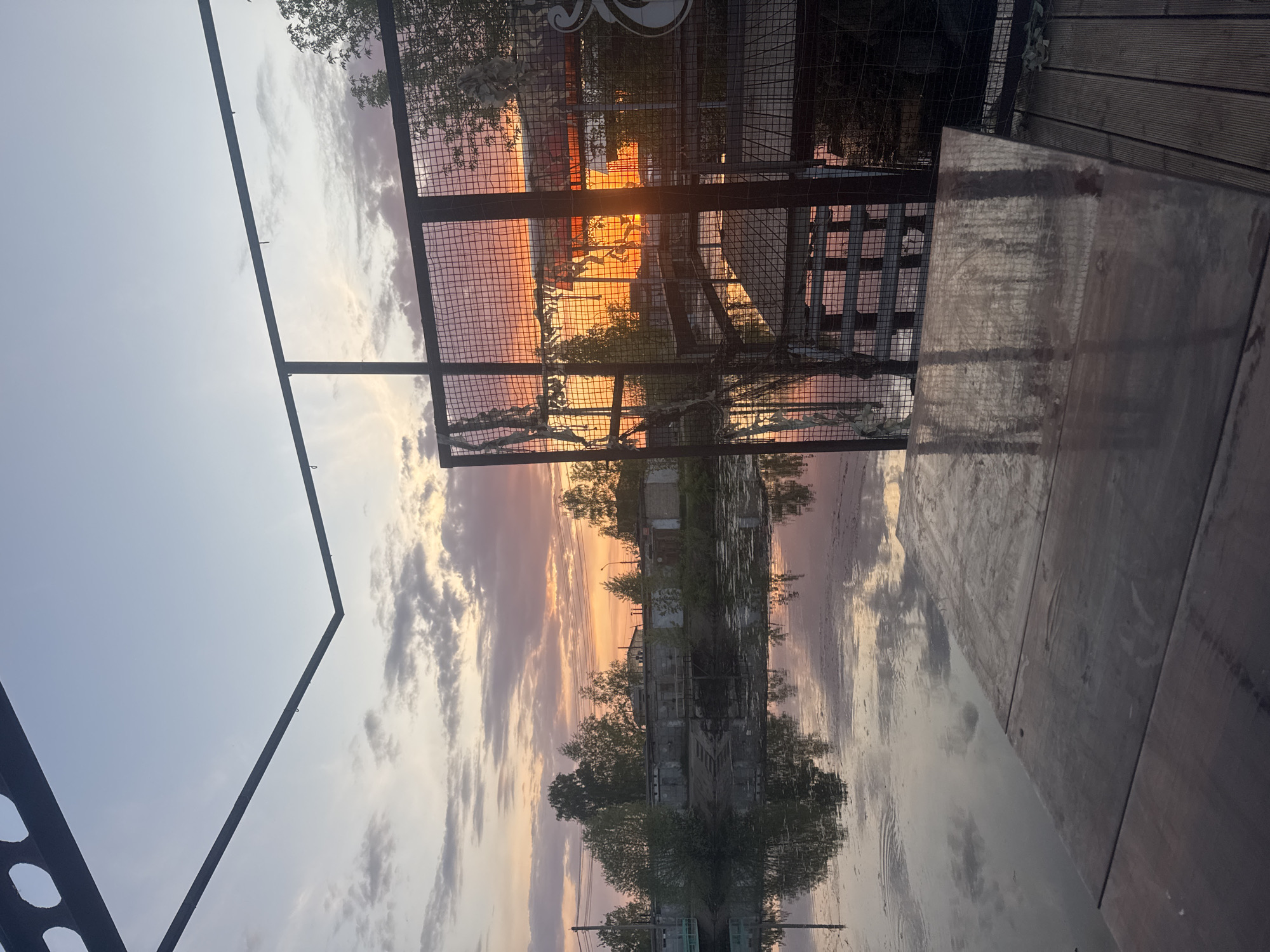 The image shows an outdoor scene in what appears to be a public space in Chaikovsky, Russia, during sunset or sunrise. A metal fence or barrier is in the foreground. Beyond the fence, there is a body of water reflecting the sky and an industrial or residential building. The sky exhibits a gradient of colors, with orange and pink hues near the horizon transitioning to lighter blues and grays. Part of a table is in the scene. The table surface is wet.