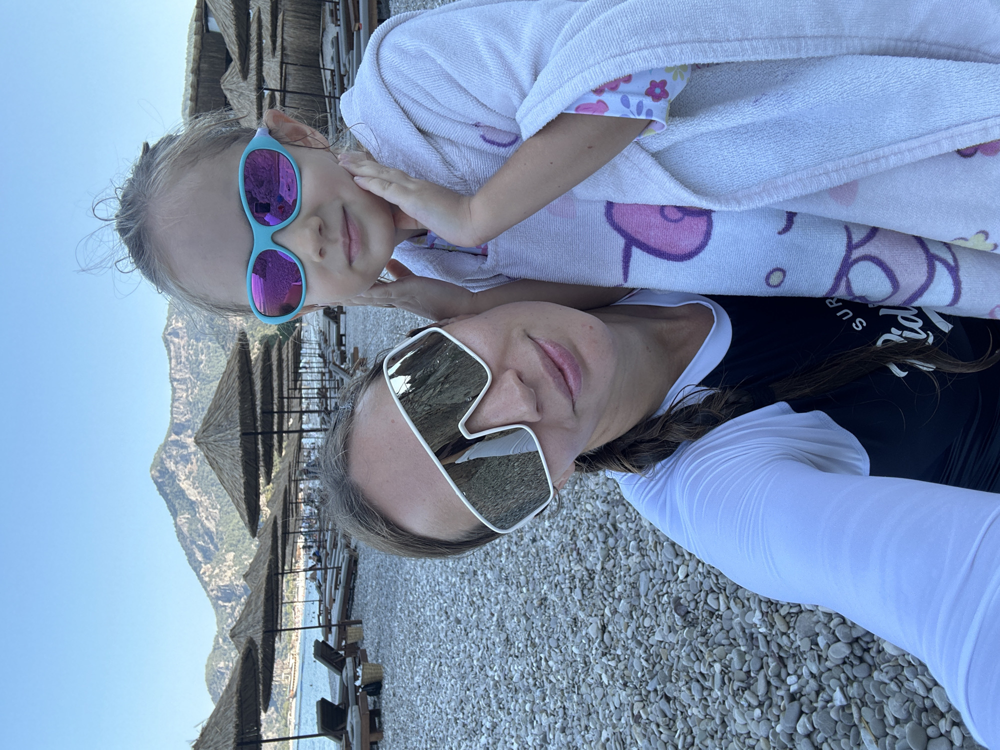 A woman and a young girl, likely mother and daughter, are taking a selfie on a pebble beach. The woman, wearing oversized white sunglasses and a white rashguard over a black swimsuit, is positioned on the left side of the frame, her arm around the girl. The girl, on the right, is wearing bright blue sunglasses and a white hooded towel with a cartoon cat pattern. Her hands are resting on her cheeks, and she is looking directly at the camera.
The background reveals a well-organized beach with rows of straw umbrellas and lounge chairs. The sea is visible in the distance, and beyond that, a range of mountains can be seen under a clear blue sky. The lighting suggests it is daytime, likely late afternoon due to the warm tones. The beach is located in the Fethiye region of Turkey, a popular tourist destination known for its stunning coastline and mountainous backdrop.