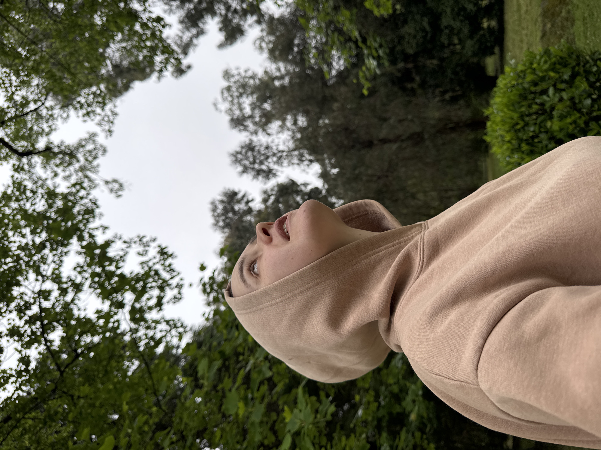 A person wearing a tan hoodie is looking up. The person's head is tilted back, and their mouth is slightly open. The background consists of trees and a cloudy sky. The location is not discernible, so a city cannot be determined.