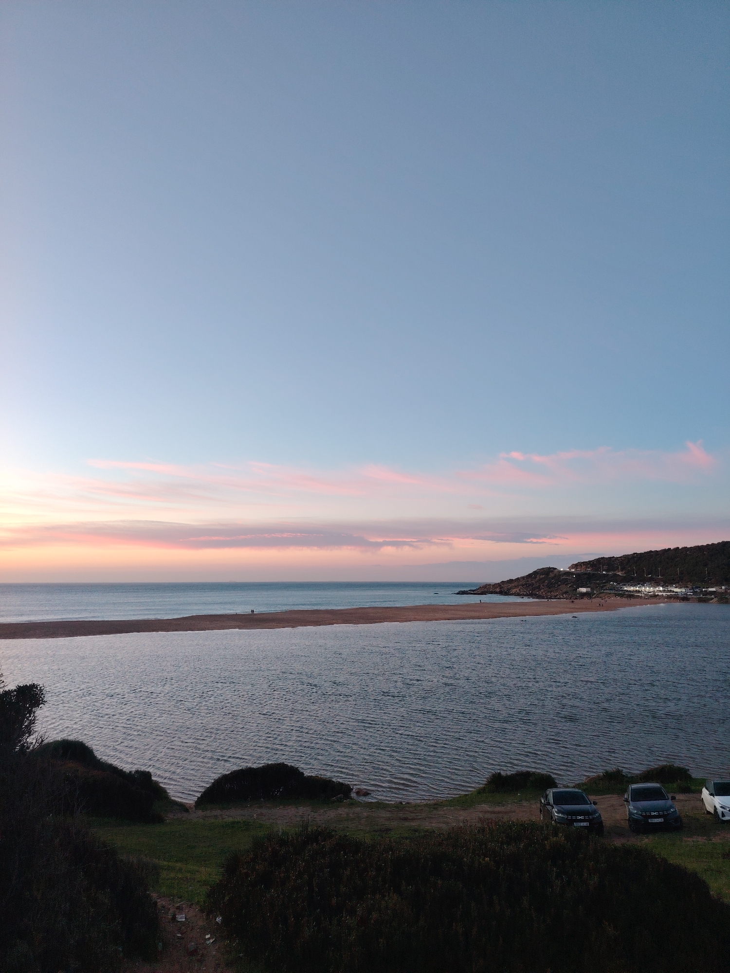 This image depicts a coastal landscape at dusk, specifically at Sidi Kacem Beach, near the mouth of Oued Tahaddart in Tanger, Morocco.
The sky occupies the upper two-thirds of the frame, transitioning from a light blue zenith to a horizon marked by horizontal bands of pink and orange stratocumulus clouds, characteristic of sunset. Below the sky, the vast expanse of the Atlantic Ocean is visible, meeting the horizon line. A long, narrow sandbar, light brown in color, separates the ocean from an inner, calm body of water, likely a lagoon or estuary. Two individuals are discernible as small silhouettes walking on this sandbar towards the left.
To the right, a rocky headland rises, covered in dark green vegetation. Scattered white buildings and illuminated structures are visible along the hillside, indicating human settlement or development. A winding road with visible lights ascends the hill. Additional small figures are present on the beach at the base of this headland.
In the foreground, a dark, elevated landmass covered in dense, low-lying dark green and brown shrubbery slopes downwards towards the inner body of water. Patches of lighter green grass are interspersed. Three vehicles are parked on a dirt patch near the water's edge on the right side of the foreground: two dark-colored SUVs/crossovers and one partially visible white car. The water in the lagoon exhibits minor ripples.