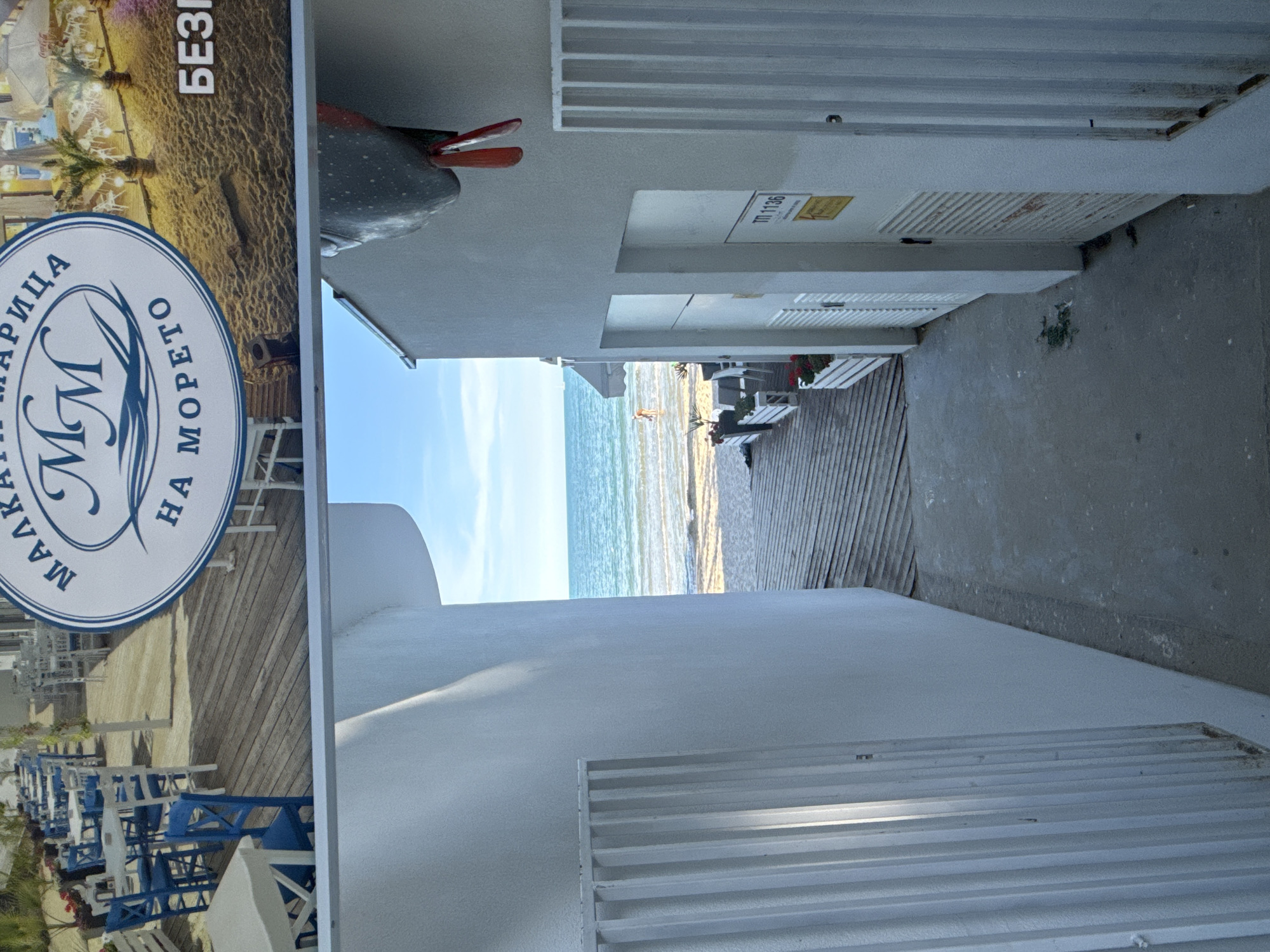 The image depicts a scene in Varna, Bulgaria, likely at a beachside establishment. The primary focus is on an outdoor seating area with numerous tables and chairs arranged on a wooden deck, suggesting a cafe or restaurant. The blue and white color scheme of the furniture is prominent. In the background, the sea is visible under a bright sky, indicating it is daytime and the weather is clear.
To the left, a large sign is partially visible. It features a circular logo with the letters "MM" in the center, surrounded by Cyrillic text. The text reads "МАРИЦА НА МОРЕТО" which translates to "Maritsa on the Sea." Above the logo, there is more Cyrillic text, "БЕЗПЛАТНО" meaning "Free."
Further to the right, the architecture of a building is visible. It has a modern, minimalist design with white walls and horizontal louvers on some sections. A utility box or panel on the building has a label with the text "ТП 1136" and a yellow warning sticker.
The overall impression is a pleasant, sunny day at a seaside resort, with an emphasis on outdoor dining or relaxation. There are no visible people in the immediate foreground, but a distant figure can be seen on the beach near the water. The scene conveys a sense of leisure and vacation.