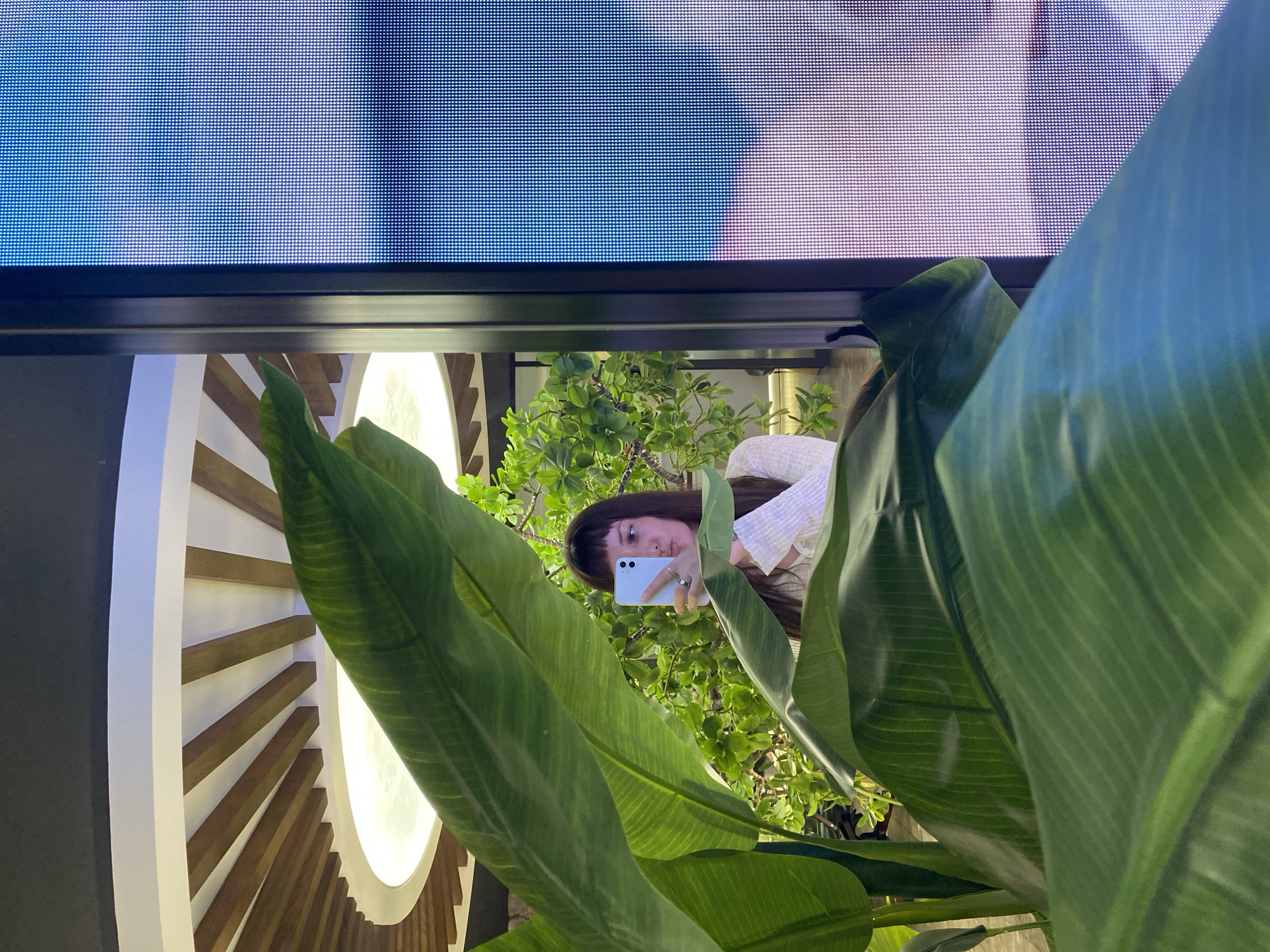 A young woman with bangs and dark hair is taking a mirror selfie with her smartphone. She is wearing a light-colored, patterned long-sleeved shirt and a ring on her right ring finger. The background reveals a modern interior with a circular ceiling feature with wooden slats and a bright light. Large, lush green leaves from a plant are in the foreground, partially obscuring the woman and the interior. To the right of the woman, a large, pixelated digital screen displays a blue and white abstract image, possibly a sky or clouds. The overall scene suggests an indoor contemporary setting, likely a public space like a mall, gallery, or a modern office. The location is Votkinsk, Russia.