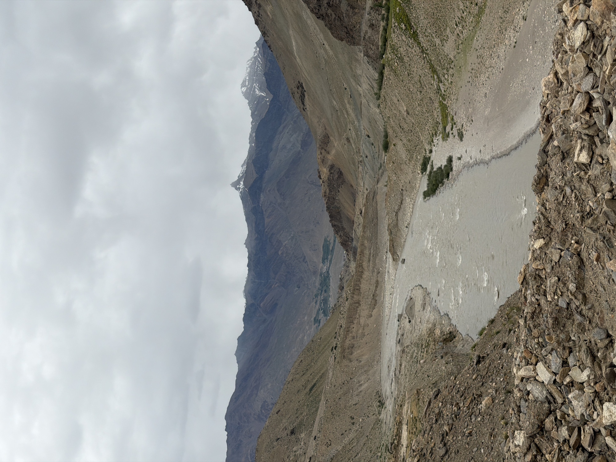 The image depicts a wide, muddy river flowing through a barren, rocky valley in Padum, India. The terrain is characterized by steep, arid slopes covered with loose rocks and sparse vegetation. In the background, imposing, layered mountains rise under a cloudy, overcast sky. The riverbed is wide and appears to be a mix of sand, gravel, and rocks. There is no visible human activity or structures in the immediate vicinity of the river, though a small settlement or village can be faintly seen nestled in the valley in the distance. The overall impression is one of a remote, rugged, and dramatic natural landscape.