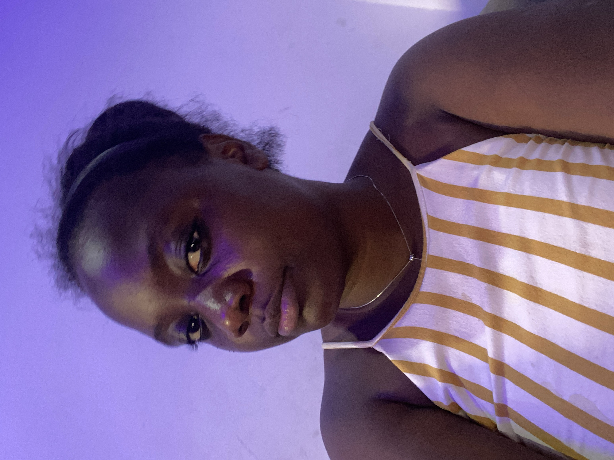 A close-up portrait shows an individual with dark skin, likely a woman, looking directly into the camera. The image is rotated 90 degrees counter-clockwise, framing the person's head and upper torso. The subject's dark hair is pulled back from the face. They are wearing a sleeveless top with white and yellow or gold horizontal stripes and a thin silver necklace with a small, circular pendant.
The lighting is a striking feature, with a dominant purple or violet hue emanating from the left side, casting a strong glow on the person's face, neck, and shoulder, as well as the background. This purple light creates distinct highlights on the nose, lips, and forehead, giving the skin a slightly luminous quality. The expression is neutral to slightly contemplative, with the eyes appearing bright and engaged with the camera.
The background is a uniform purple, suggesting an indoor setting with colored ambient lighting. There is no visible text. The setting is in Abuja, Nigeria. The image appears to be a close-up photograph or selfie, capturing the individual in a relaxed pose.