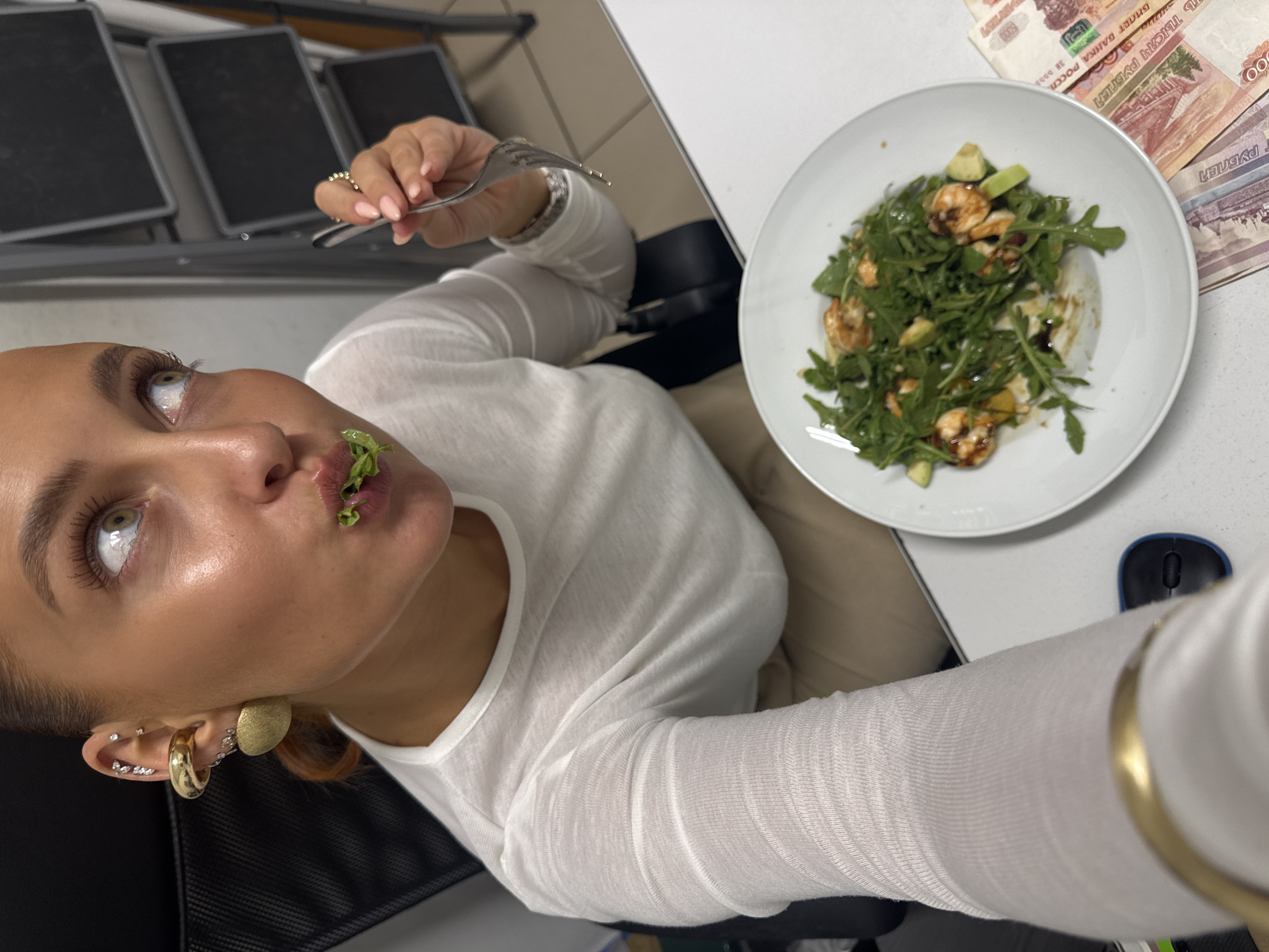 A woman is seated at a desk, eating a salad. She is wearing a long-sleeved white shirt and gold earrings. She is holding a fork with salad on it, near her mouth. Her gaze is upwards. On the desk next to the plate of salad, there are stacks of Russian rubles, specifically 5000-ruble notes. A computer mouse is visible on the desk near her right arm. Behind her there are 3 folded chairs, suggesting that this could be a working lunch. The city of Chaikovsky, Russia, is not identifiable from the image alone.
