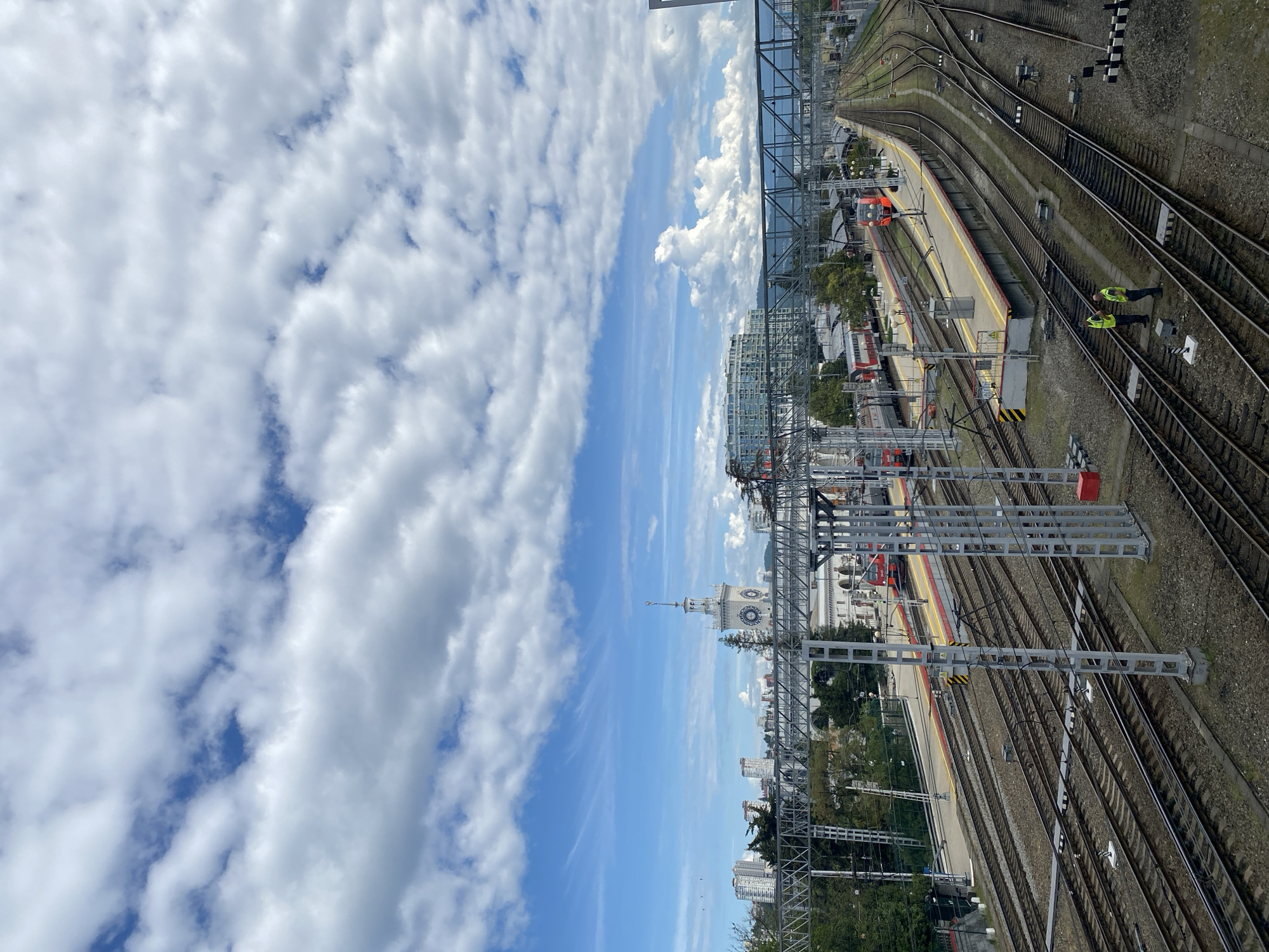 A high-angle, medium shot depicts a railway scene under a sky with scattered clouds. The image seems rotated to be viewed vertically. Railway tracks dominate the right side, with two people wearing yellow vests near the tracks. A train station platform with yellow accents can be seen. Overhead electric lines span across the tracks.
In the background, a white clock tower stands prominently amid greenery and city buildings. Further back, a modern glass building and a waterfront area are visible. This is likely a view of the Sochi railway station in Sochi, Russia, due to the distinctive clock tower and railway infrastructure.