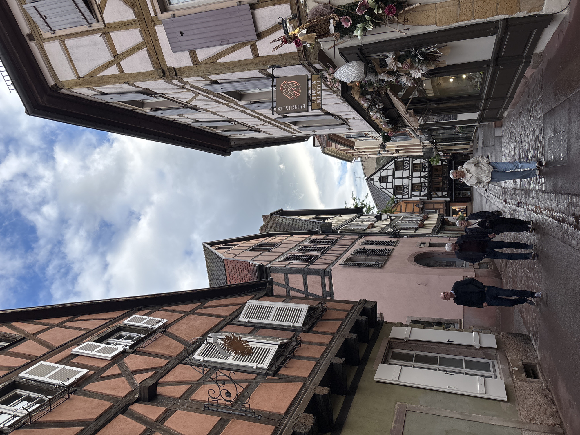 The image shows a narrow cobblestone street in Colmar, France, lined with traditional half-timbered houses. The buildings feature colorful facades, exposed wooden beams, and shuttered windows. On the right side of the street, a shop displays decorative floral arrangements and a sign that reads "EMPREINTES BIJOUX CREATION." Three people are walking down the street. Two men are walking side-by-side, and a woman is walking behind them. The sky is cloudy with patches of blue. The street appears wet, suggesting recent rain. The architecture and atmosphere are characteristic of the picturesque old town of Colmar, likely in the Petite Venise (Little Venice) district, known for its charming canals and well-preserved medieval buildings.
