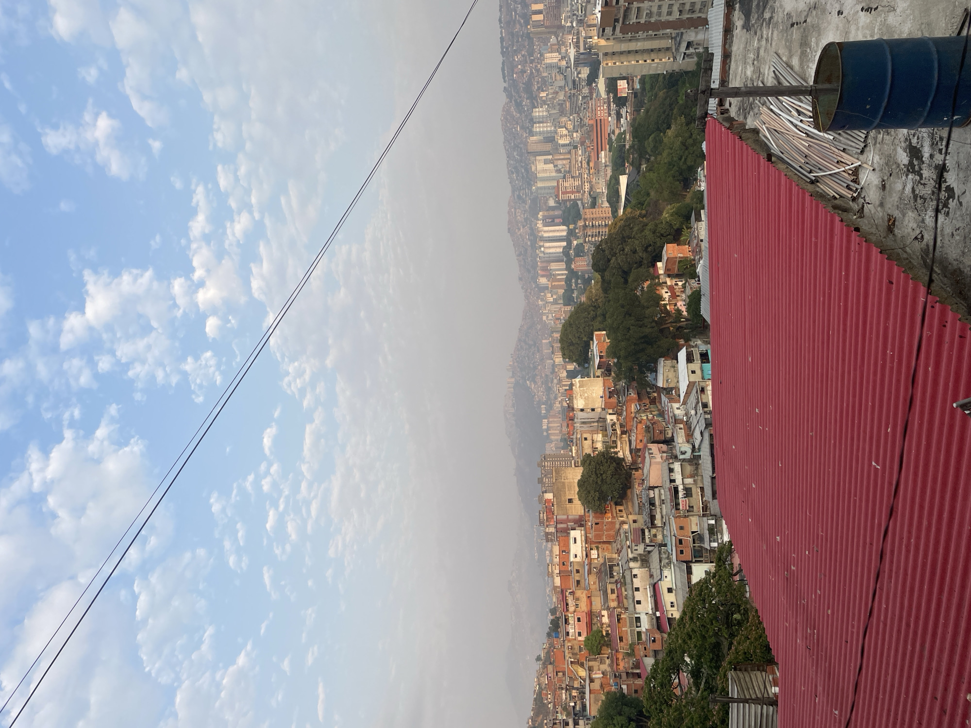 The image shows a view of Caracas, Venezuela, taken from a rooftop. The foreground features a red corrugated metal roof and a blue barrel. A pile of what appear to be fluorescent light tubes are stacked next to the barrel. The cityscape in the background is a mix of tall buildings and densely packed, smaller structures, likely representing a favela or barrio. A layer of smog or haze obscures the distant buildings. The sky is blue with scattered clouds. Power lines stretch across the sky.