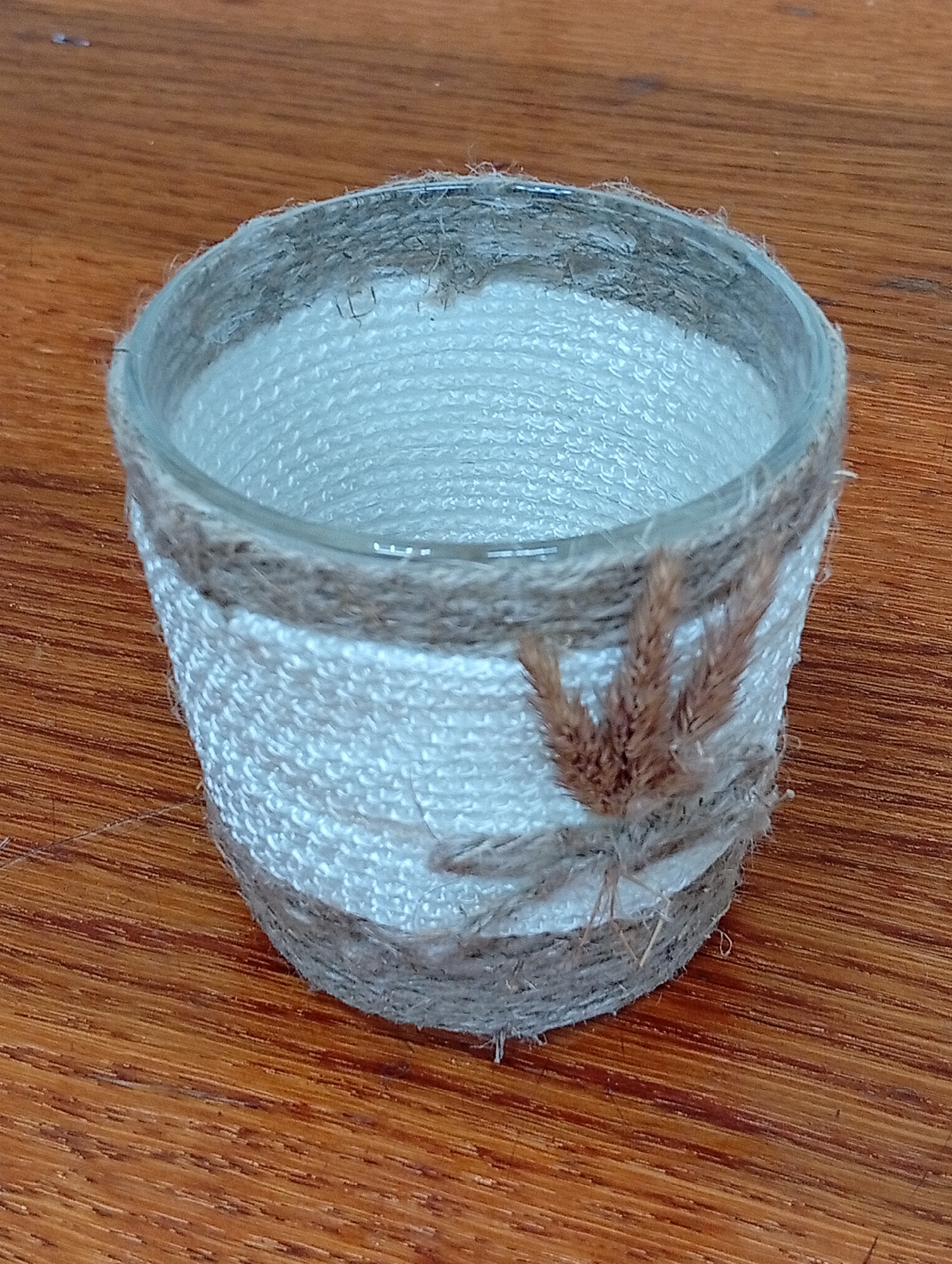 A small, cylindrical glass container, likely a repurposed jar, is decorated with twine. The twine is wrapped horizontally around the exterior of the glass. A lighter colored, textured material, possibly fabric or a thicker twine, forms the main body of the decoration, with two bands of darker, coarser twine at the top and bottom. A small bunch of dried grass or reeds is attached to the darker twine band in the foreground, serving as embellishment. The container rests on a wooden surface with a visible grain. The background is blurred, suggesting the image was taken indoors, possibly on a table. There is no indication of the location being in Крыўлянскі сельскі Савет, Belarus.