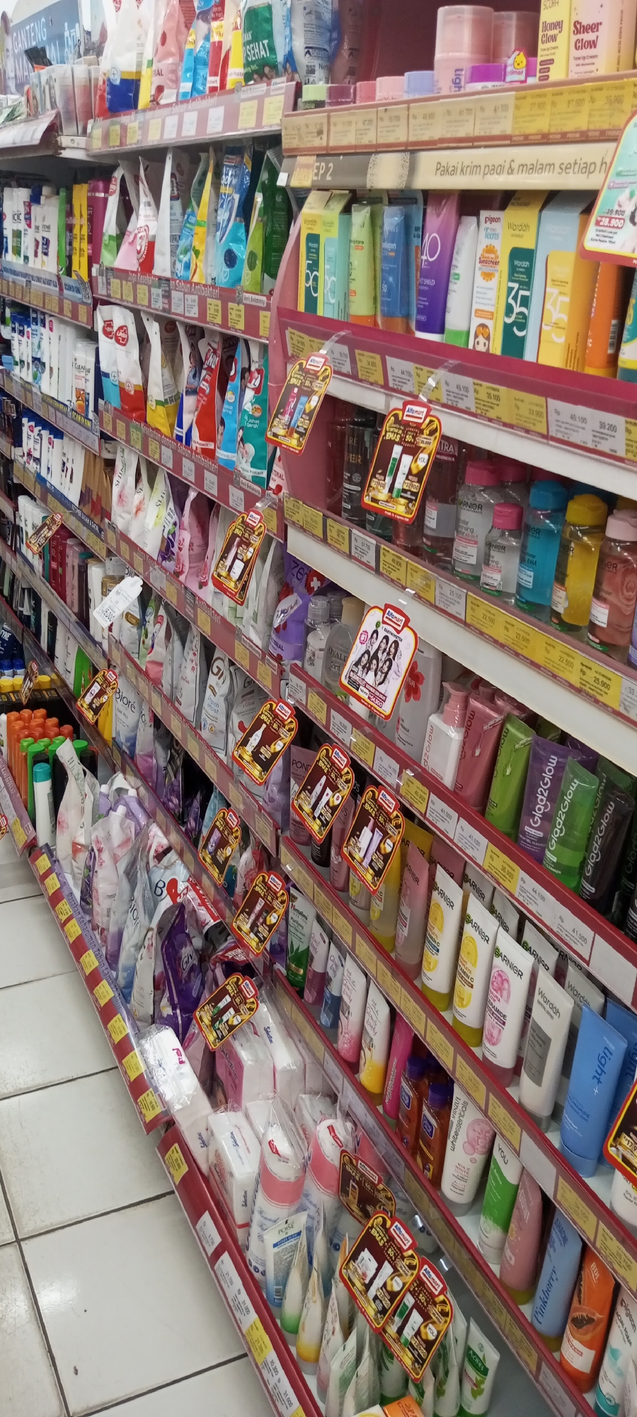 The image shows an aisle of a brightly lit convenience store, likely an Alfamart, in Setu, Indonesia. The shelves are packed with a wide variety of personal care and beauty products. Products are displayed in rows, with many items in brightly colored packaging. Some shelves feature promotional tags, indicating discounts or special offers. The perspective is slightly angled, looking down the aisle and showcasing a dense arrangement of merchandise. The flooring appears to be white tile. The overall impression is one of a well-stocked retail environment focused on fast-moving consumer goods.