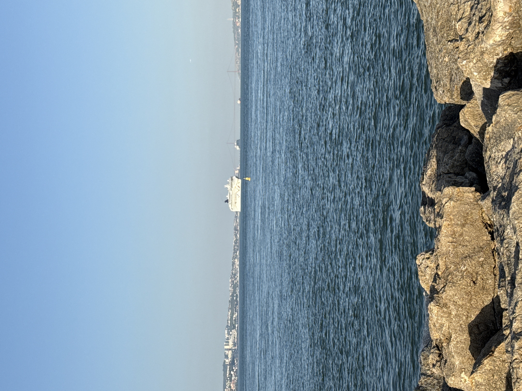 A wide-angle, eye-level, vertical shot captures a bright, sunny day with a clear, pale blue sky. The foreground is dominated by large, light-brown rocks with deep shadows, suggesting a rocky shoreline. Beyond the rocks, a vast expanse of dark blue, slightly rippled water stretches to the horizon. In the mid-ground, a white cruise ship is visible on the water, and further in the distance, a cityscape with numerous buildings lines the shore. A large red suspension bridge spans the water in the far distance. The scene appears to be the Tagus River, likely near Oeiras, Portugal, given the presence of the suspension bridge, which resembles the 25 de Abril Bridge. No people are clearly visible, and the focus is on the natural and man-made elements of the waterfront.