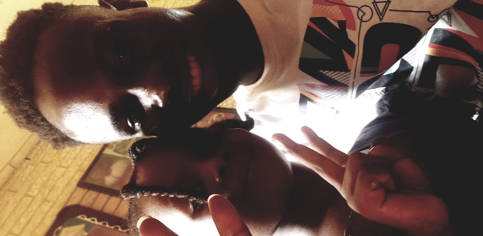 This close-up, warm-toned selfie depicts two individuals, an adult male and a young female, situated indoors in the city of Narayi, Nigeria. The male, positioned on the right, has dark skin, short curly hair, and is wearing a light-colored t-shirt with a multi-colored geometric pattern across the chest. He is smiling, revealing teeth. His face exhibits strong highlights and shadows, with light illuminating his forehead and left eye, while his right cheek and eye are partially obscured by shadow. The female, positioned on the left, has dark skin and braided hair. Her face is largely shadowed, with her left eye partially visible. Her left hand is raised, making a V-sign with her index and middle fingers, and her thumb shows white nail polish. The background behind them appears to be an interior space, with a textured, light brown wall visible on the left and a framed object, possibly a picture or mirror, further in the background.