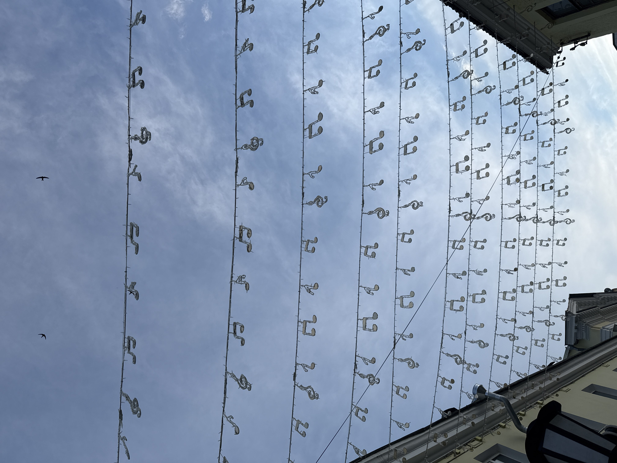 The image presents a daytime scene looking upwards towards a partly cloudy sky, featuring an extensive overhead display of decorative lights shaped like musical notes. Numerous rows of these light-colored decorations, primarily depicting treble clefs and eighth notes, are suspended by wires across the view.
The sky, a bright blue with scattered white clouds, fills most of the frame. Two small, dark birds are visible in flight against the sky on the left side, one more prominent than the other.
On the right side, the upper section of a building is visible, including a roofline with an overhang, a gutter, and a section of a wall, possibly light-colored. The decorations appear to extend from fixtures associated with this building.
The scene suggests a festive or themed public area in Заволжье, Russia, adorned with musical motifs. The clear daylight indicates good weather. No visible text is present in the image.