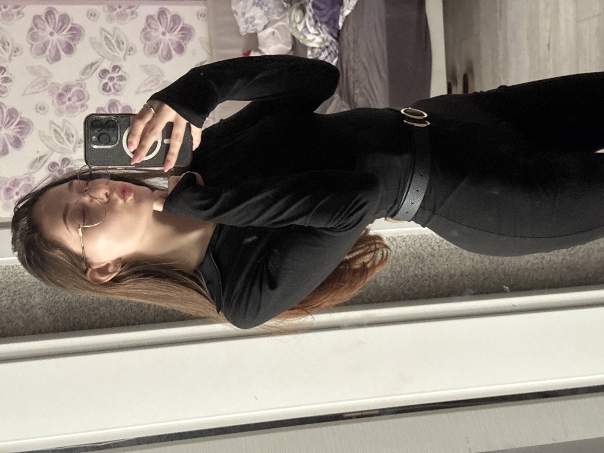 An indoor mirror selfie captures a young woman posing with a playful "kiss face" expression. She has long, straight brown hair and is wearing round, wire-frame glasses. Her left hand, adorned with a silver ring and light pink manicured nails, holds a dark-colored smartphone, which has a glittery clear case featuring a prominent circular MagSafe-style ring.
She is dressed in a long-sleeved black top and form-fitting black pants, cinched at the waist by a black belt with a distinctive gold-colored double-ring buckle.
The background, visible as a reflection behind her in the mirror, features a wall adorned with light purple floral wallpaper on the left side. To the right of the wallpaper, there appears to be a bed or sofa with rumpled grey and purple bedding or clothes. The floor in the foreground, where she is standing, is covered in light grey carpet, with a white baseboard or trim running along the bottom edge of the mirror. The setting suggests a personal living space, such as a bedroom or dressing area. The image does not contain any visible text. The location context provided is Sofia, Bulgaria, though this is not visually discernible within the image.