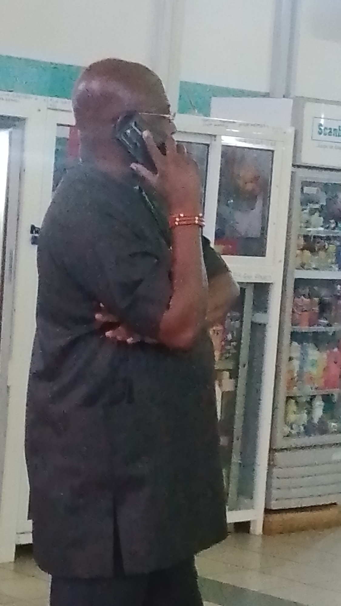 A man with a bald head, wearing a black traditional outfit and eyeglasses, is talking on a mobile phone. He has two red beaded bracelets on his right wrist. He appears to be standing in front of a display refrigerator filled with various items, possibly in a store or a public establishment in Orerokpe, Nigeria. The refrigerator has a white frame and clear glass doors. Behind the man, a reflection in a window shows another person, partially obscured. The environment appears to be indoors, with light-colored walls and flooring.