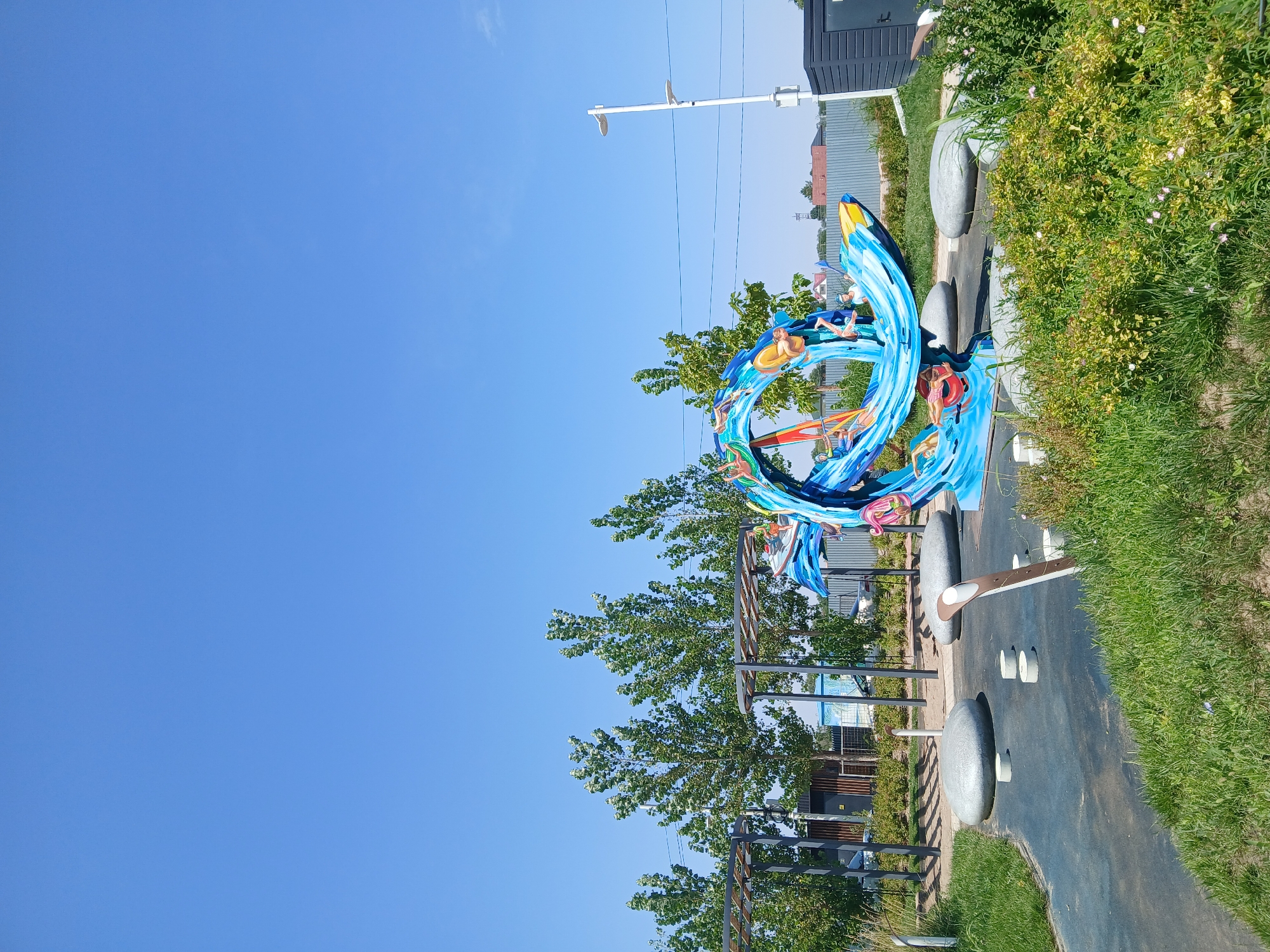 The image depicts an outdoor public space with a vibrant, blue, wave-like sculpture adorned with colorful figures of people engaged in water sports. The sculpture appears to be made of painted metal or fiberglass and is situated on a dark blue, rubberized surface, likely a playground or park area. Scattered around the sculpture are large, smooth, gray, pebble-like seating elements and smaller circular concrete steps.
In the background, there are rows of tall, leafy green trees, and behind them, a gray metal fence encloses an area with some buildings and structures. A white lamppost with two lights stands to the right of the sculpture, and a dark gray rectangular structure, possibly a utility box or small building, is also visible. The sky is a clear, bright blue, indicating a sunny day.
The scene is located in the city of Qonaev, Kazakhstan. The overall impression is of a modern, recreational urban area designed for public enjoyment. There are no visible people in the image, but the sculpture's design suggests activity and a connection to water-based recreation. The ground is covered with grass and low-lying greenery in some areas, particularly in the foreground and to the right.