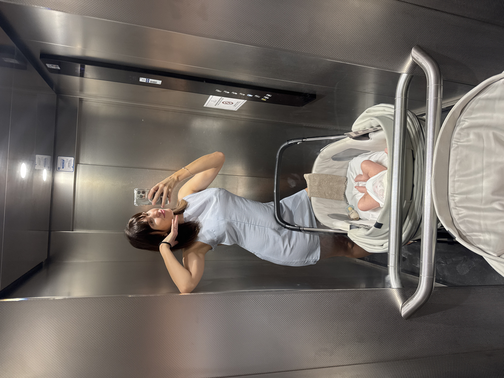 The image is a mirror selfie taken inside an elevator. A woman with dark hair and bangs is posing with her left arm bent and hand touching her head, while holding a smartphone in her right hand. She is wearing a light blue and white striped sleeveless dress. In the foreground, a baby stroller is positioned, with a newborn baby visible inside, wearing a diaper and what appears to be a light pink onesie. The baby's legs and feet are visible. The elevator interior features textured metallic walls, a black panel with elevator buttons, and a brushed metal handrail. The floor is dark. There are no external elements visible to confirm a location in Isola delle Femmine, Italy.