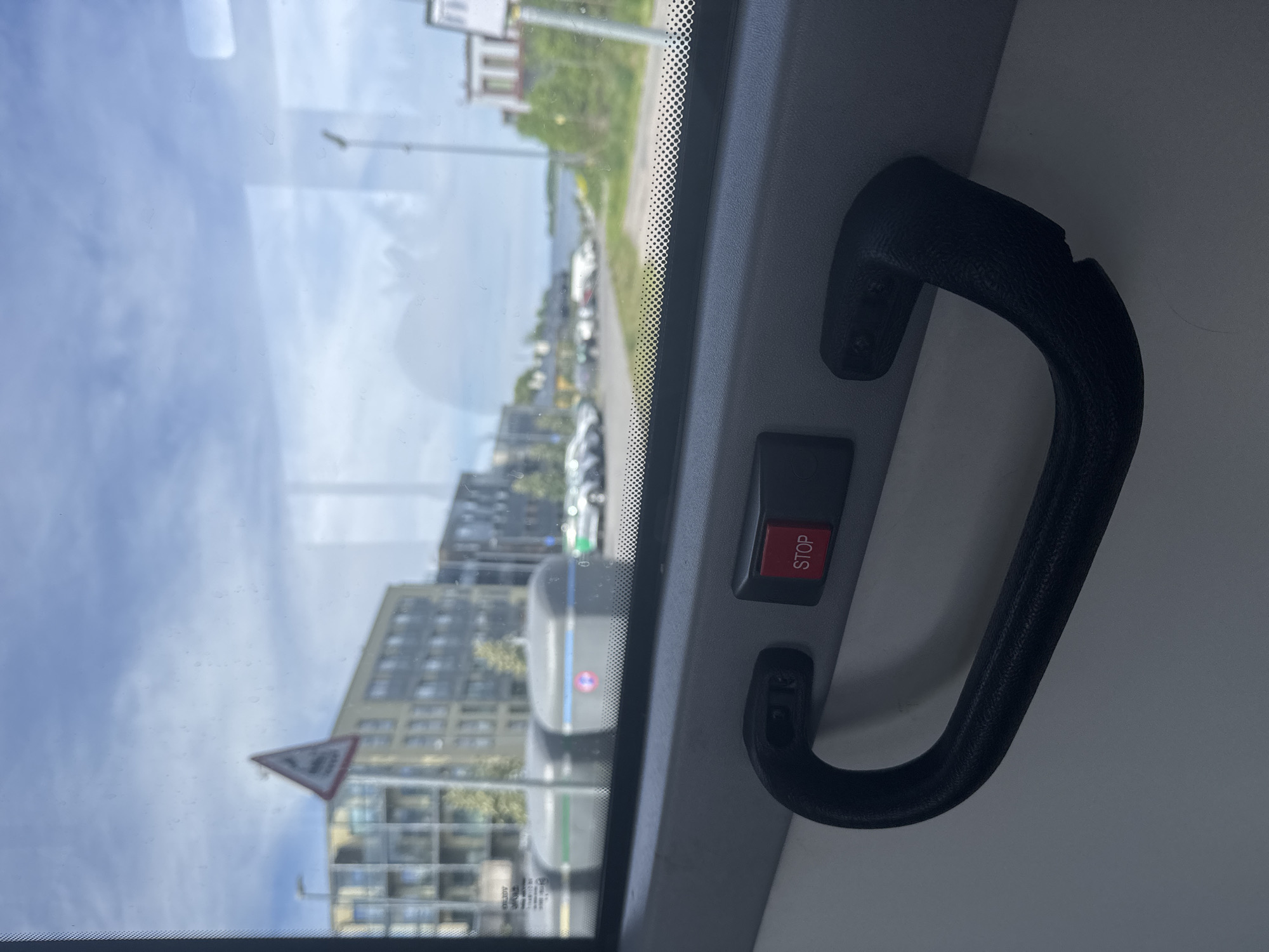 The image captures a view from inside a bus, looking out through the window. In the foreground, a black bus handle is prominently displayed, along with a red "STOP" button. The window looks out onto an urban scene with modern apartment buildings, parking areas with cars, and what appears to be a body of water in the distance. A triangular warning sign with an image of a pedestrian is visible on a pole outside. The sky is partly cloudy. The scene is consistent with the urban environment of Tallinn, Estonia, particularly in areas with mixed-use development, such as residential buildings near parking and waterfront access. There are no people clearly visible in the image.