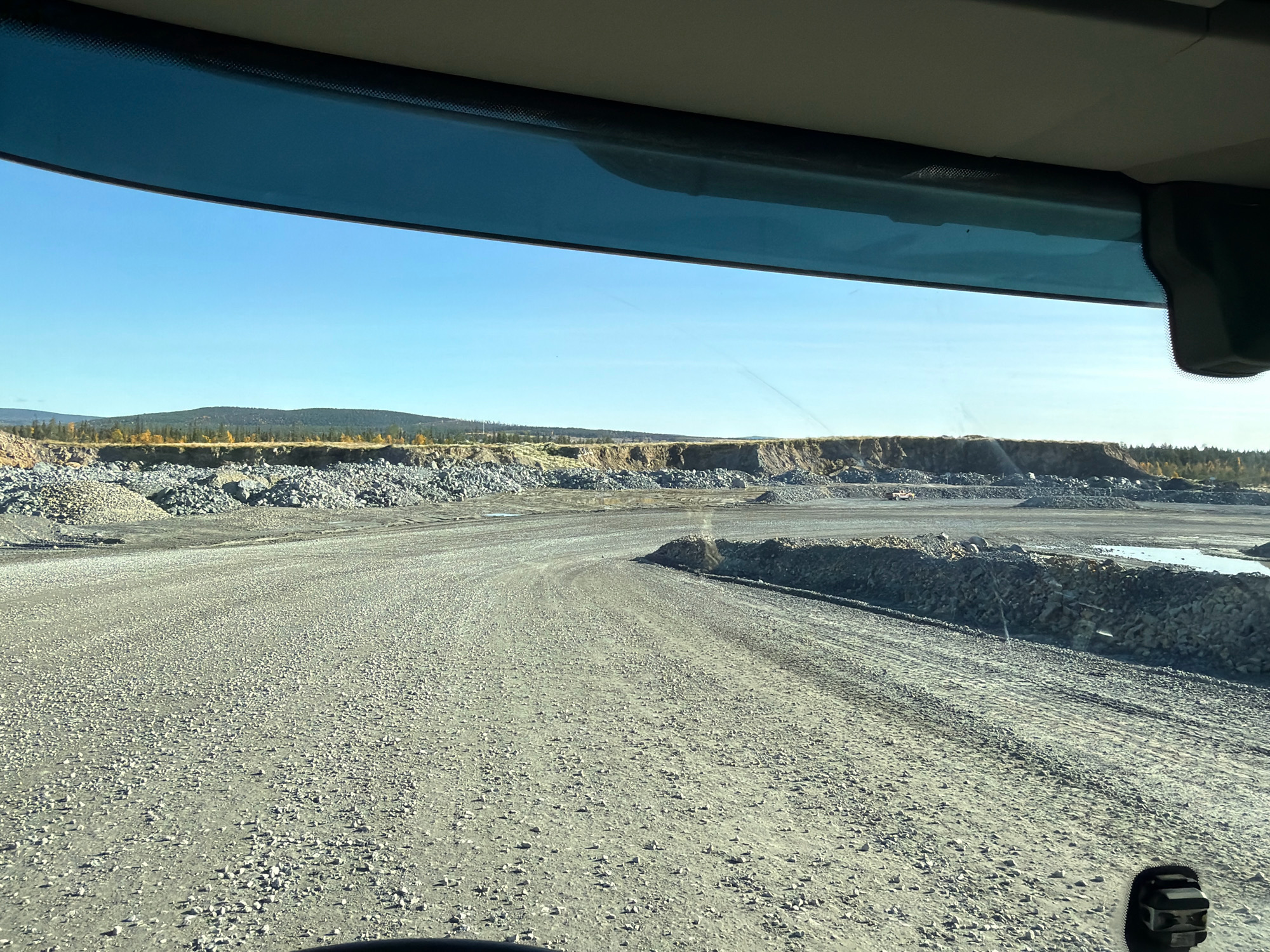 The image was taken inside a vehicle, looking out through the windshield at a quarry under a clear blue sky. The foreground is dominated by a gravel road. In the middle ground, there are piles of rocks and a water-filled area. The background features a rugged landscape with more rock piles and a distant treeline. The image is from a quarry in Gällivare, Sweden, likely part of the LKAB mine operations. There are no people visible in the image.