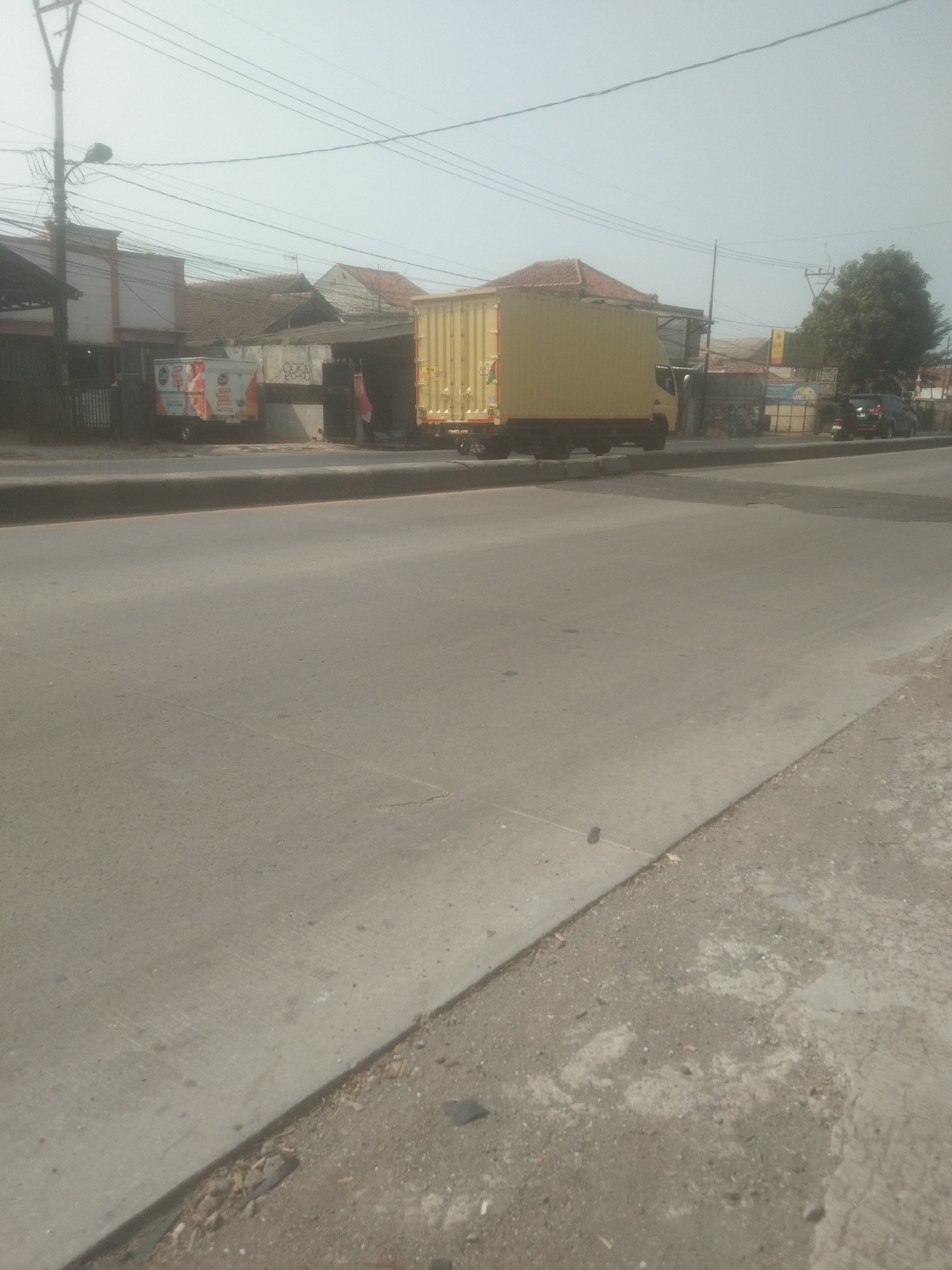 A wide shot captures a street scene with a concrete road in the foreground and buildings and vehicles on the right side of the road. A utility pole stands on the left, with numerous wires crisscrossing the sky. A bright yellow box truck is parked on the right side of the road, facing away from the camera. Next to it, a smaller white truck with graphics is also parked. Further to the right, a dark SUV is visible, and behind it, a green tree stands out against the pale sky. The buildings in the background appear to be a mix of commercial structures and residential homes with tiled roofs. The overall scene suggests an urban environment in Subang, Indonesia, possibly during daylight hours.