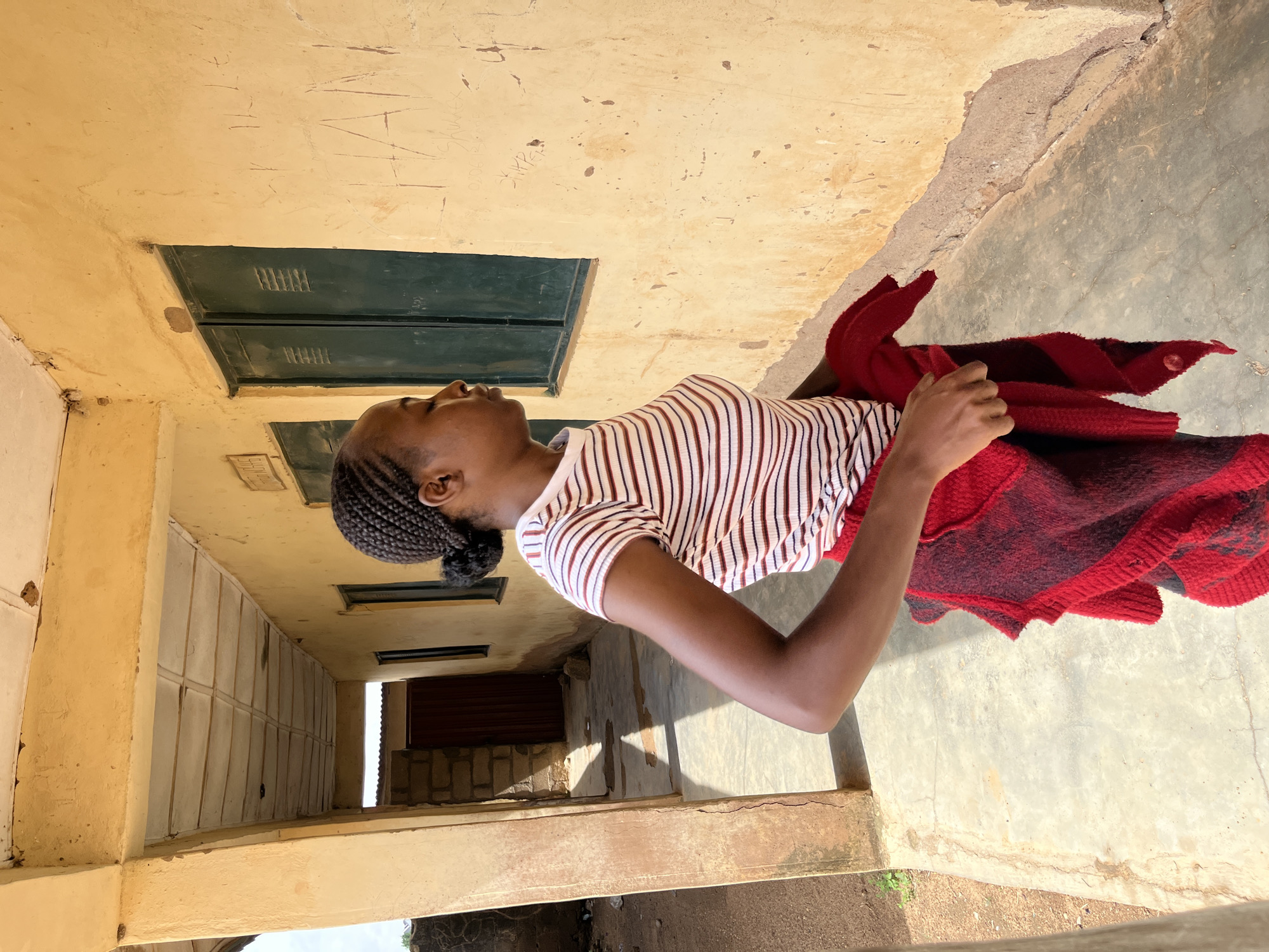 A young Black woman with braided hair, wearing a horizontally striped short-sleeved shirt and holding a red and black patterned wrap, stands in a covered walkway with a yellow stucco wall and green-shuttered windows. The walkway has a concrete floor with light and shadow patterns. The wall has graffiti, including the word "HAN" scratched into it. In the background, a dark brown door is visible, and beyond that, a corrugated metal roof. The environment appears to be part of a building in Bukuru, Nigeria. The image is taken from a medium distance, focusing on the woman and the adjacent wall.