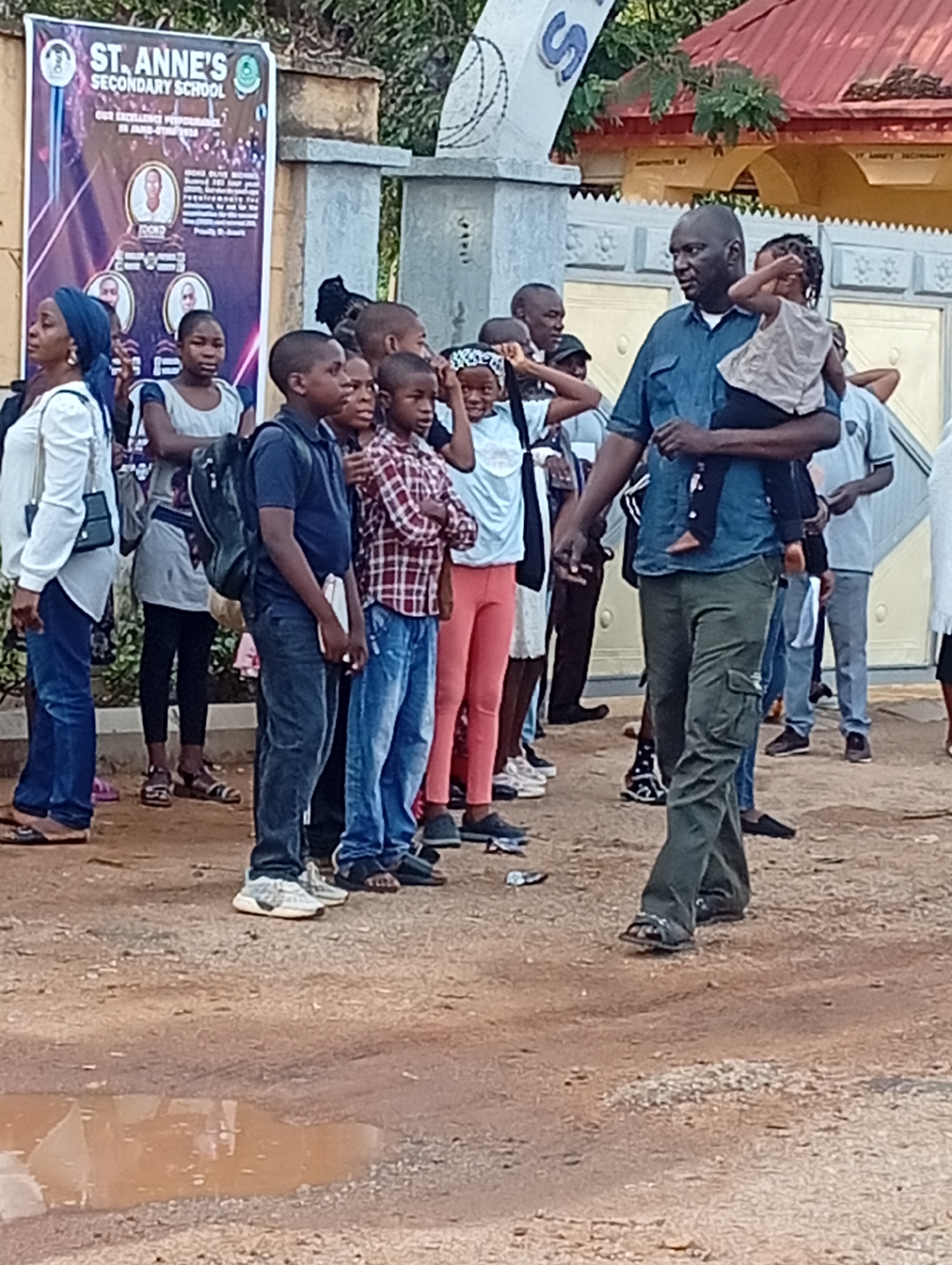 The image captures a group of individuals standing near a banner for St. Anne's Secondary School in Barden Yana, Nigeria. Several students, some in uniform and others in casual clothing, are present. An adult male carries a young child. The background includes a building with a red roof and a gated entrance, along with some foliage. The ground is a dirt surface with a puddle.