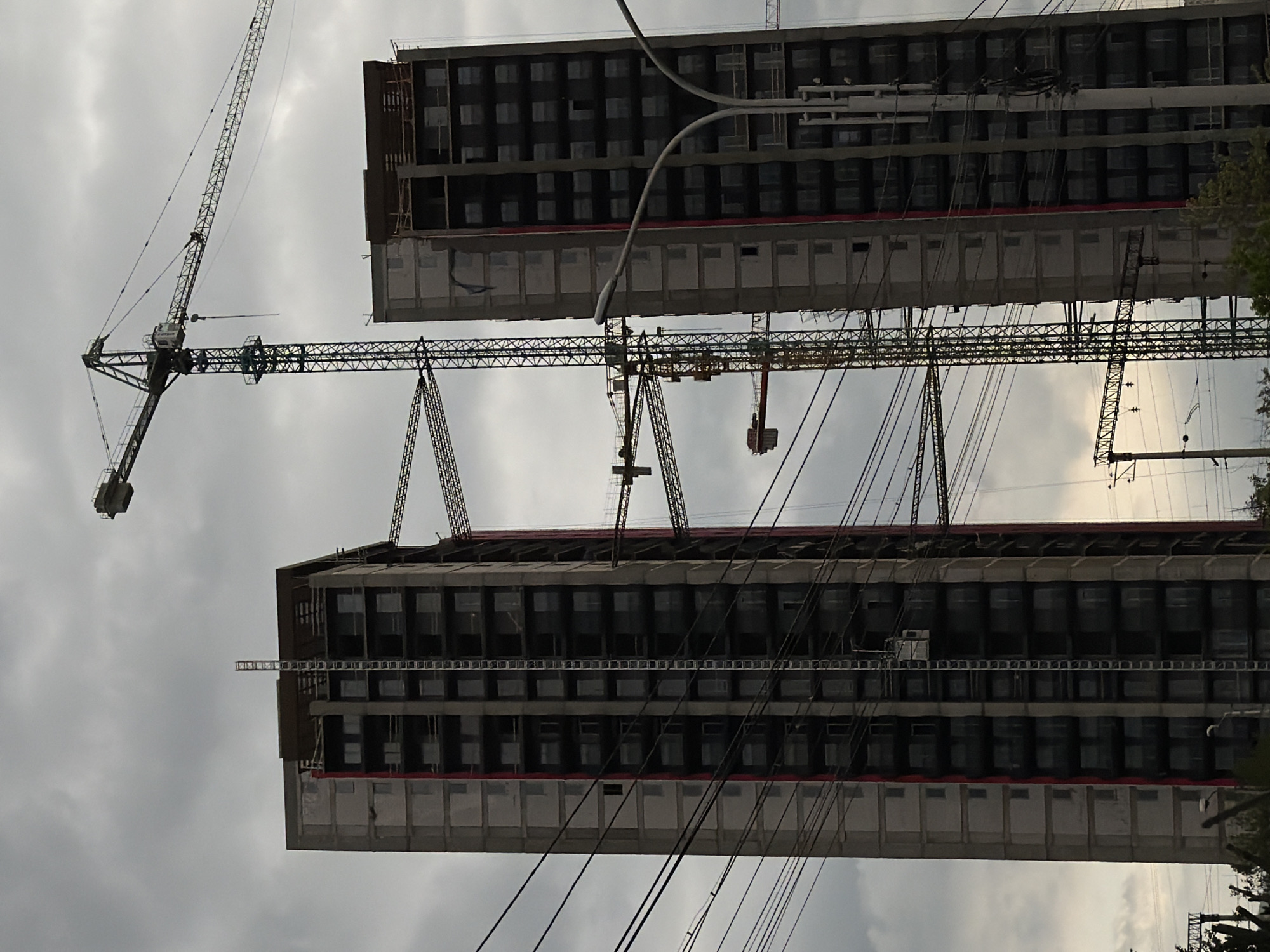 The image depicts two partially constructed high-rise buildings in Tbilisi, Georgia. Both buildings appear to have a regular grid of windows and a horizontal red stripe near the top. Numerous cranes are actively involved in the construction. Power lines are visible in the foreground and middle ground. The sky is overcast.