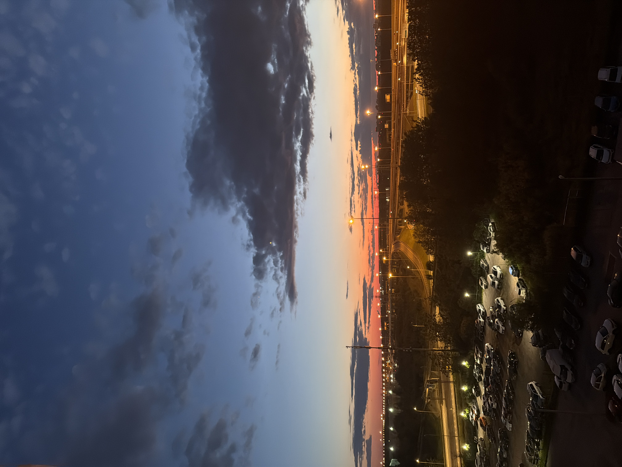 A dusk aerial view captures a parking lot filled with an array of cars, illuminated by streetlights against a backdrop of a highway and a vibrant sunset sky. The sky transitions from a deep blue at the top to hues of orange and red near the horizon, with scattered clouds adding texture. In the foreground, a dark mass of trees partially obscures the scene. The parking lot, situated in the lower half of the image, is densely packed with vehicles of various makes and models, many of which are white, black, or silver. Beyond the parking lot, a multi-lane highway curves through the landscape, with cars visible on its elevated sections, their headlights and taillights casting streaks of light. Numerous streetlights line the highway and the surrounding area, providing illumination. In the distance, the faint glow of city lights suggests an urban environment. No people are visible in the image.
