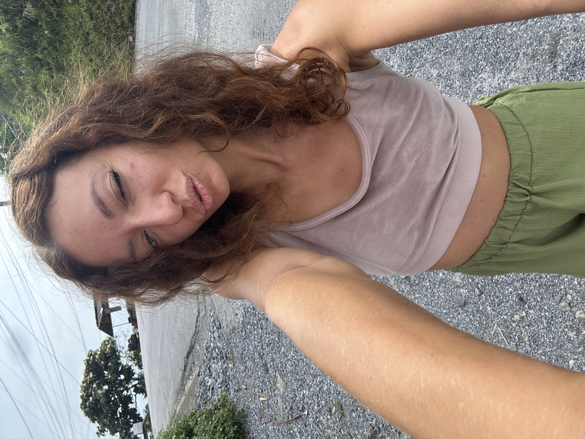 A woman is taking a selfie outdoors. She is fair-skinned with curly auburn hair, wearing a pale pink cropped top and olive green pants. She is winking at the camera and pouting her lips. The background shows a street with a gravel surface. There is a building and trees in the distance. Based on the details in the image, including the style of architecture and vegetation, the location is consistent with the city of Tam Son, Vietnam.