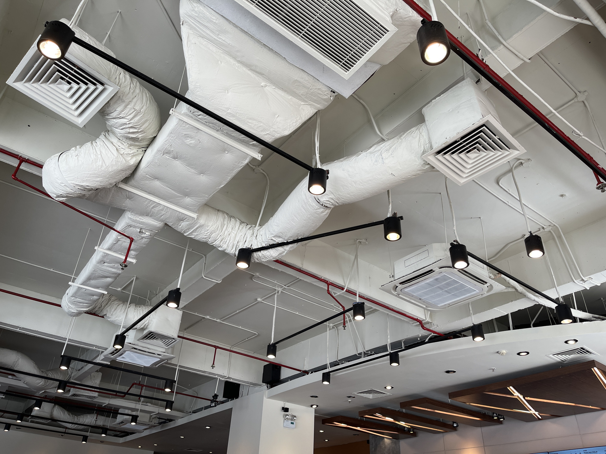 This image captures the interior ceiling of a commercial establishment, likely a cafe or modern office space, in Đà Nẵng, Vietnam. The design showcases an exposed, industrial-chic aesthetic with various utilitarian elements prominently displayed against a white ceiling.
The most striking features are the extensive network of white, insulated HVAC (heating, ventilation, and air conditioning) ducts that snake across the ceiling. These ducts vary in shape, some appearing as large rectangular channels and others as flexible, convoluted tubes. Interspersed among them are numerous air diffusers and grilles, including several large square grilles with concentric louvers and rectangular units.
A modern black track lighting system runs throughout the space, featuring multiple cylindrical black spotlight fixtures that cast a bright, focused glow. Red and white electrical conduits or pipes (possibly fire suppression lines) are also visible, running alongside and beneath the ducts and lighting tracks, contributing to the exposed utility theme. Several white, ceiling-mounted air conditioning units, including a cassette-type unit, are integrated into the ceiling.
In the lower part of the image, the ceiling transitions to a more finished appearance with recessed lighting and a decorative wooden panel structure that incorporates linear illumination. A white emergency light fixture with unidentifiable text is mounted on a wall. Below the decorative ceiling, a display screen or menu board shows some blurred, illegible text. No people or specific activities are present in the static architectural shot. The time of day is not discernible due to the indoor setting and artificial lighting.