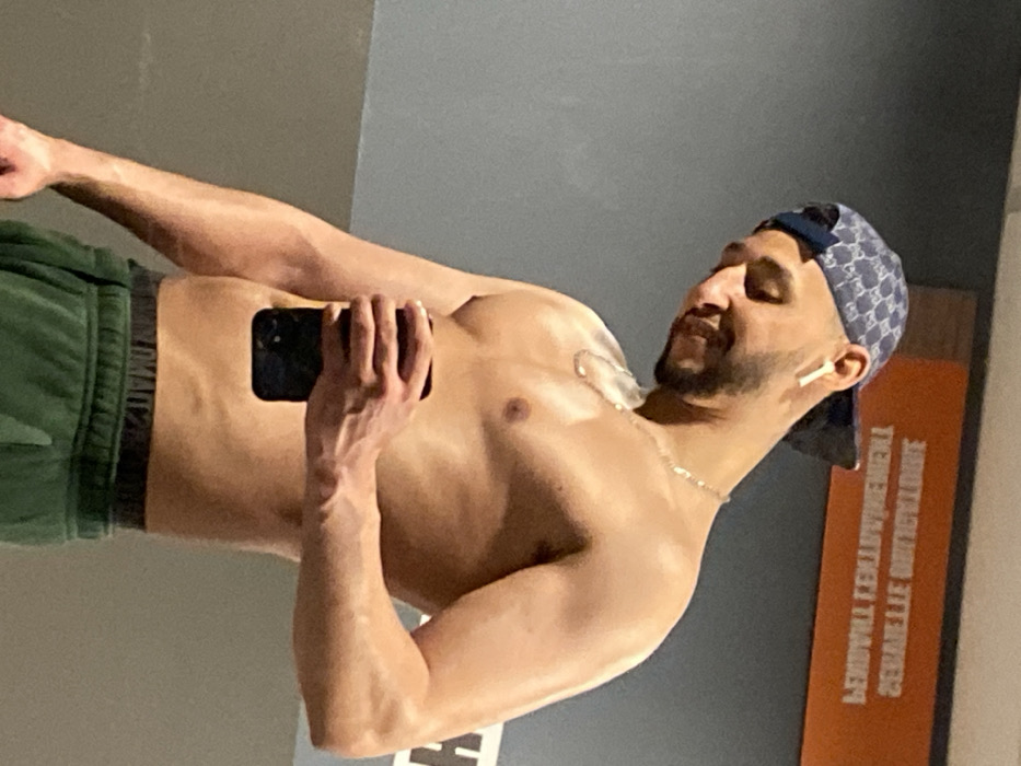 A shirtless male individual is depicted from the mid-chest upwards, observed in what appears to be a mirror reflection. He faces slightly right, his head tilted downwards, exhibiting a subtle smile. He wears a blue patterned baseball cap backward, a white wireless earbud in his right ear, and a thin silver chain necklace. His physique is muscular, and his skin appears moist. He holds a black smartphone vertically in his left hand, obscuring a portion of his lower torso. Green athletic pants with a black elastic waistband are visible below his waist. The background consists of a grey wall and an orange panel bearing inverted white French text that reads "SERVIETTE OBLIGATOIRE" (Towel obligatory) and "ENTRAINEMENT" (Training). This scene is consistent with an indoor fitness facility, specifically located in Hœnheim, France, based on the linguistic context of the background text.