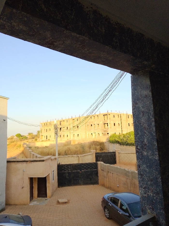 A high-angle view from Bar Gada, Nigeria, depicts an outdoor scene featuring multiple structures and a dry environment under a clear blue sky. In the immediate foreground, a dark, textured concrete beam extends horizontally across the top, supported by a similarly textured concrete column on the right, indicating the vantage point is from within a building or overhang.
Below, the ground area is paved with reddish-brown interlocking pavers. A dark gray sedan is parked in the lower right, with the rear half visible. Another vehicle, partially obscured, is present in the lower left. On the left side of the paved area, a single-story, light brown building with a dark brown door and a single window stands. A square concrete slab is positioned on the ground in front of this building. A dark, double-leaf gate and a light-colored block wall enclose the paved area.
Beyond this enclosed space, the mid-ground reveals an expanse of dry, yellowish-brown vegetation and sandy earth. A tall, light-colored utility pole with multiple black electrical wires strung across it stands centrally. In the background, a large, multi-story building, appearing to be under construction, is prominent. It features numerous empty rectangular window openings and exposed rebar protruding from its roofline. A patch of green foliage is visible to the right of this building. Another light-colored wall or building section is visible on the far left mid-ground, with a distant, dome-like structure on a slight rise behind it. No individuals or active interactions are observed in the scene.