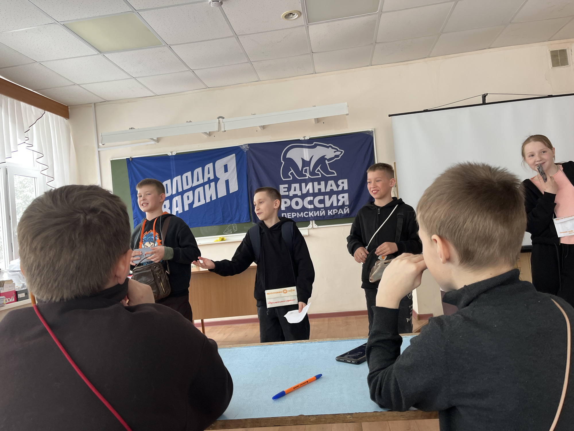 The image shows a classroom setting in Chaikovsky, Russia, indicated by the banners "Молодая Гвардия" (Young Guard) and "Единая Россия. Пермский край" (United Russia. Perm Territory). Three young boys are standing and addressing an audience sitting at desks. The boy in the center holds a paper with the words "Хорошево Новое" (Khoroshevo Novoe). The girl holds a microphone. An orange pen is seen on the table in front of the two people at desks.