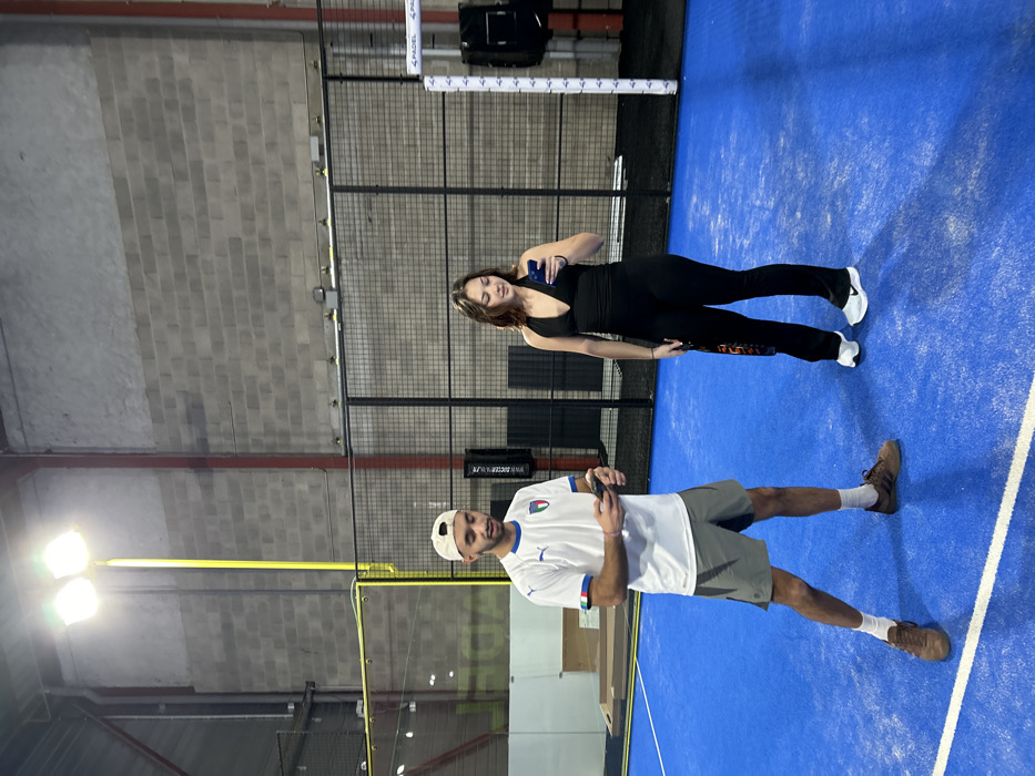 Two individuals, a man and a woman, are captured posing on an indoor padel court. The bright blue court surface features white boundary lines, with tall black mesh fencing and gray concrete block walls enclosing the playing area. Overhead, artificial lighting illuminates the scene.
The woman stands on the right, wearing a black tank top and black fitted leggings, paired with white athletic shoes. She holds a dark smartphone in her right hand, positioned near her chest. A padel racket with an orange and black pattern is partially visible by her left leg. She has long brown hair.
On the left, the man is dressed in a white short-sleeved jersey featuring the Italian flag emblem and the word "ITALIA" on the left chest. He wears gray shorts, white socks, and brown athletic shoes. A white baseball cap is worn backward on his head. He is also holding a dark smartphone in his left hand.
Both subjects appear to be standing still, possibly for a photograph, rather than actively playing. In the background, part of the padel net system is visible, with white posts displaying the word "PADEL" repeatedly. A black sign further in the background shows partially legible text "WWW.SOCCERBLU...". The setting is an indoor sports facility, specifically a padel court located in Eckbolsheim, France. The presence of artificial lights indicates an indoor setting where the time of day is not a factor.