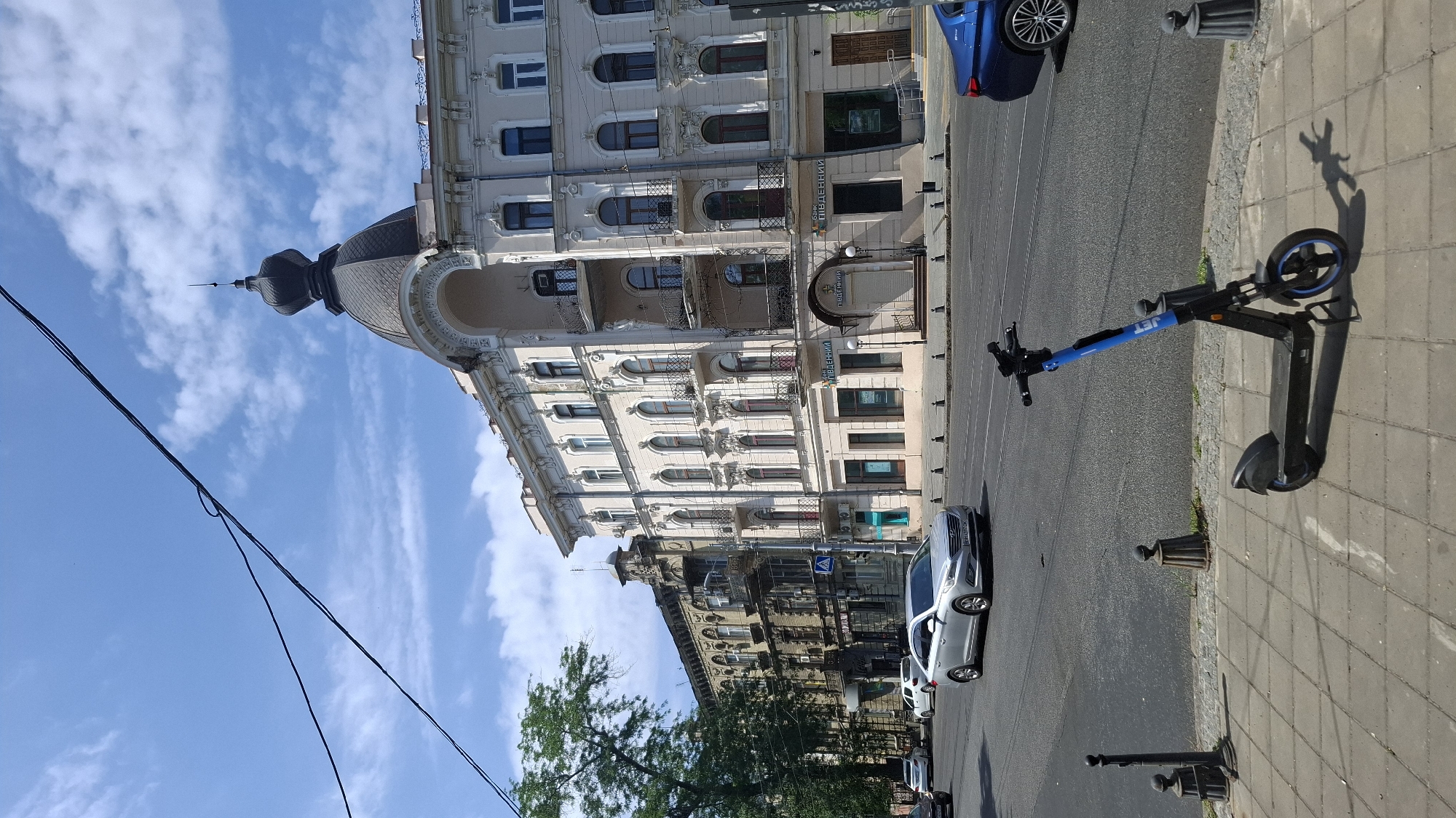 A street-level, full shot captures a bright, sunny day with a clear blue sky and scattered clouds. In the foreground, a black electric scooter with blue accents stands parked on a grey, tiled sidewalk. The scooter casts a distinct shadow on the ground. Behind the sidewalk, a multi-lane asphalt road is visible, with a silver sedan and a blue car in motion. The silver sedan is in the left lane, and the blue car is in the right lane, partially visible.
The background features a row of historic, multi-story buildings lining the street. The building on the right is ornate, with a prominent dome topped by a spire. It is painted a light cream color and features numerous arched windows, balconies with wrought-iron railings, and detailed architectural trim. The building to its left is darker, appearing older and more weathered, with a similar architectural style of windows and balconies. Trees with lush green foliage are visible on the far left side of the image. Electrical wires stretch across the top portion of the frame. The scene appears to be in an urban environment.