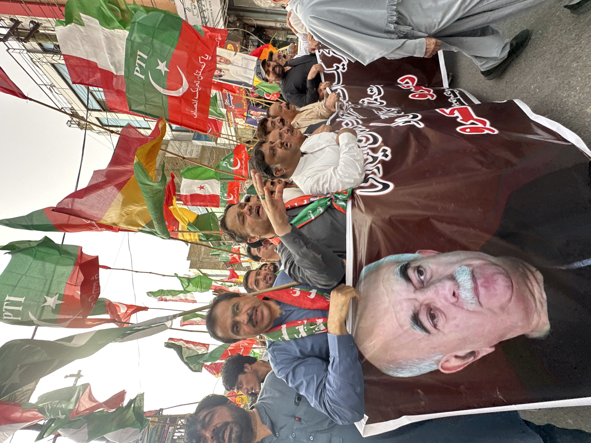 A group of men are gathered in Quetta City Tehsil, Pakistan, holding various flags and a banner. The flags are predominantly green and white, some bearing the insignia of the Pakistan Tehreek-e-Insaf (PTI) party, which includes a crescent moon and star. Several individuals are wearing traditional Pakistani clothing and political scarves. In the foreground, a large banner displays the portrait of an older man with a mustache and white hair. The men appear to be participating in a political rally or procession. The background shows a street scene with buildings and overhead electrical wires, indicating an urban environment. The sky is overcast.