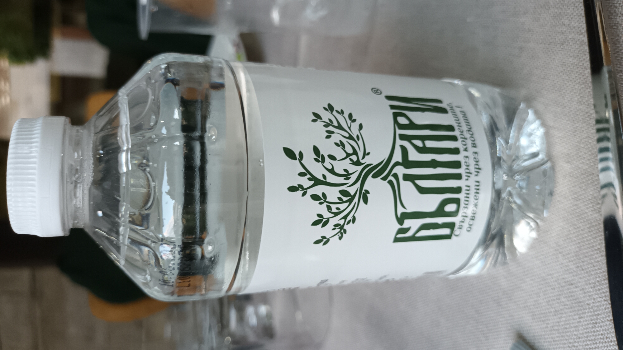 The image shows a clear plastic bottle of water. The bottle has a white cap and a label with a green tree logo and text in Bulgarian. The text on the label reads "БЪЛГАРИ" and below that "Свързани чрез корените, освежени чрез водата!". The bottle is sitting on a light-colored, textured surface, likely a tablecloth, with a metallic object partially visible in the foreground. In the background, blurry figures and objects suggest an indoor or semi-outdoor setting. There is no specific indication of the city of Razlog, Bulgaria within the image itself.