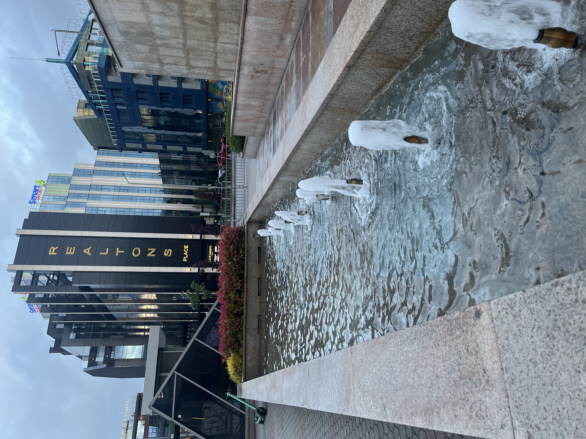 This image captures a scene in Sofia, Bulgaria, showing a long, narrow water fountain with several water jets spraying into it. The fountain is made of light-colored stone. In the background, there are several modern buildings. One building is labeled "REALTONS PLACE" in vertical, gold lettering. Adjacent to this building are flowers. A scooter is parked nearby. The sky is overcast.