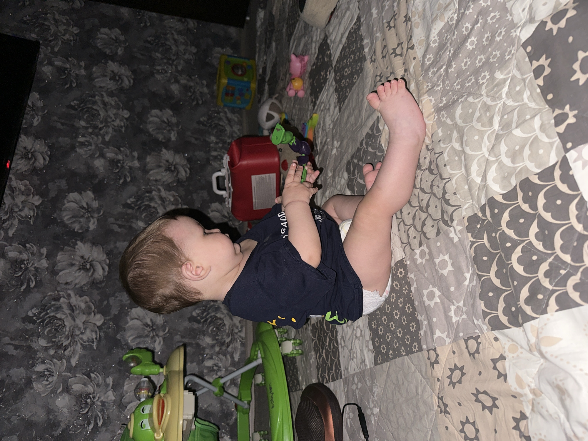 A light-skinned baby with brown hair, wearing a dark blue t-shirt and a diaper, is sitting on a multicolored patchwork quilt. The baby is turned to the right and appears to be playing with a purple and green toy. In the background, there is a green baby walker with a duck-shaped toy, a red medical bag, and a multi-colored toy cube. The wall behind the baby is covered in floral patterned wallpaper. The scene is indoors.