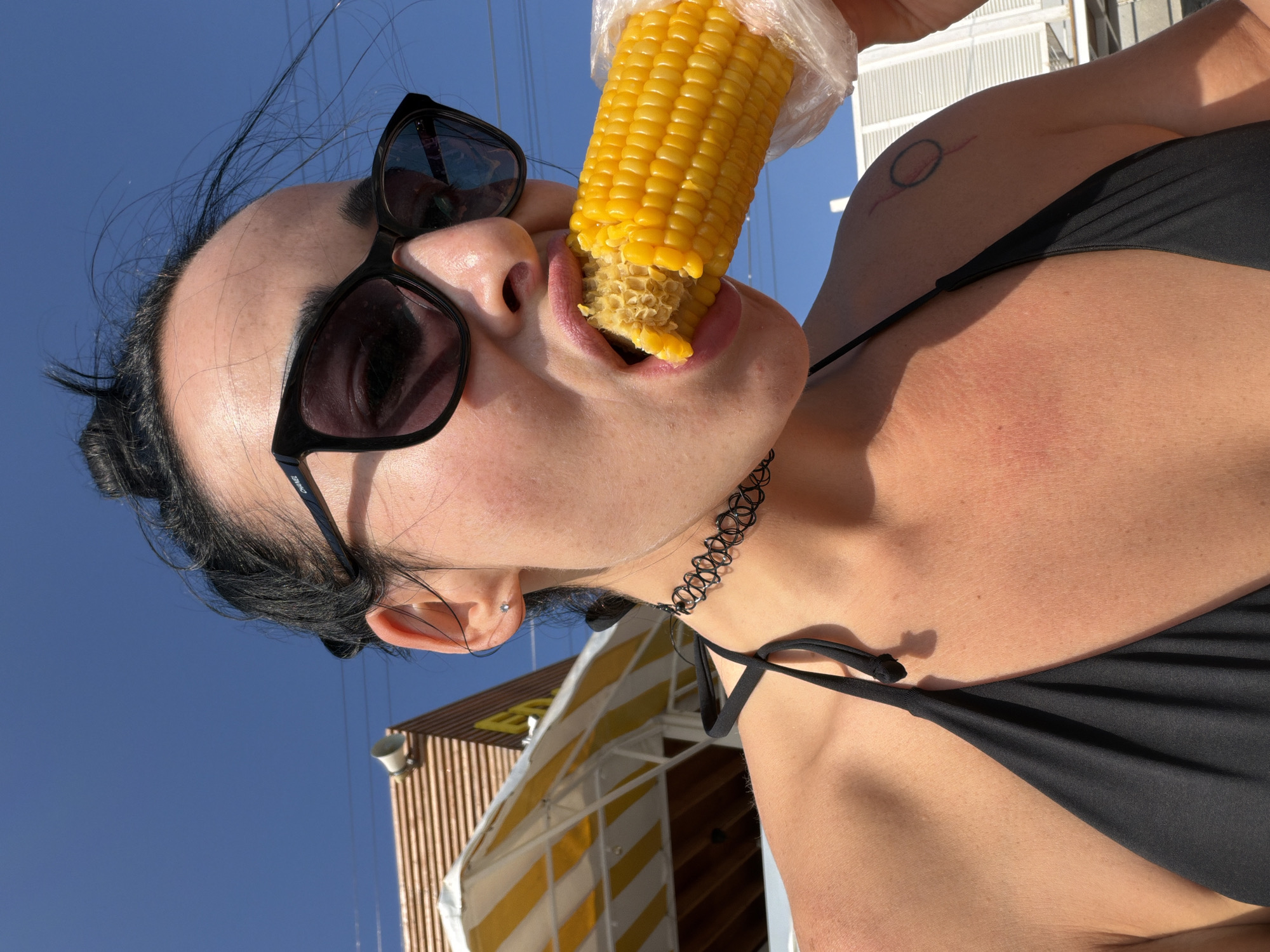 A woman with dark hair pulled up in a bun wears black sunglasses and a black bikini top. She has a black choker necklace around her neck. The woman is eating a piece of corn on the cob, which is wrapped in plastic. Her left shoulder has a circular tattoo. She is outdoors with a bright blue sky in the background. To her left, part of a yellow and white striped awning and a building are visible. The overall scene suggests a sunny day, possibly at a beach or outdoor market in Sochi, Russia.