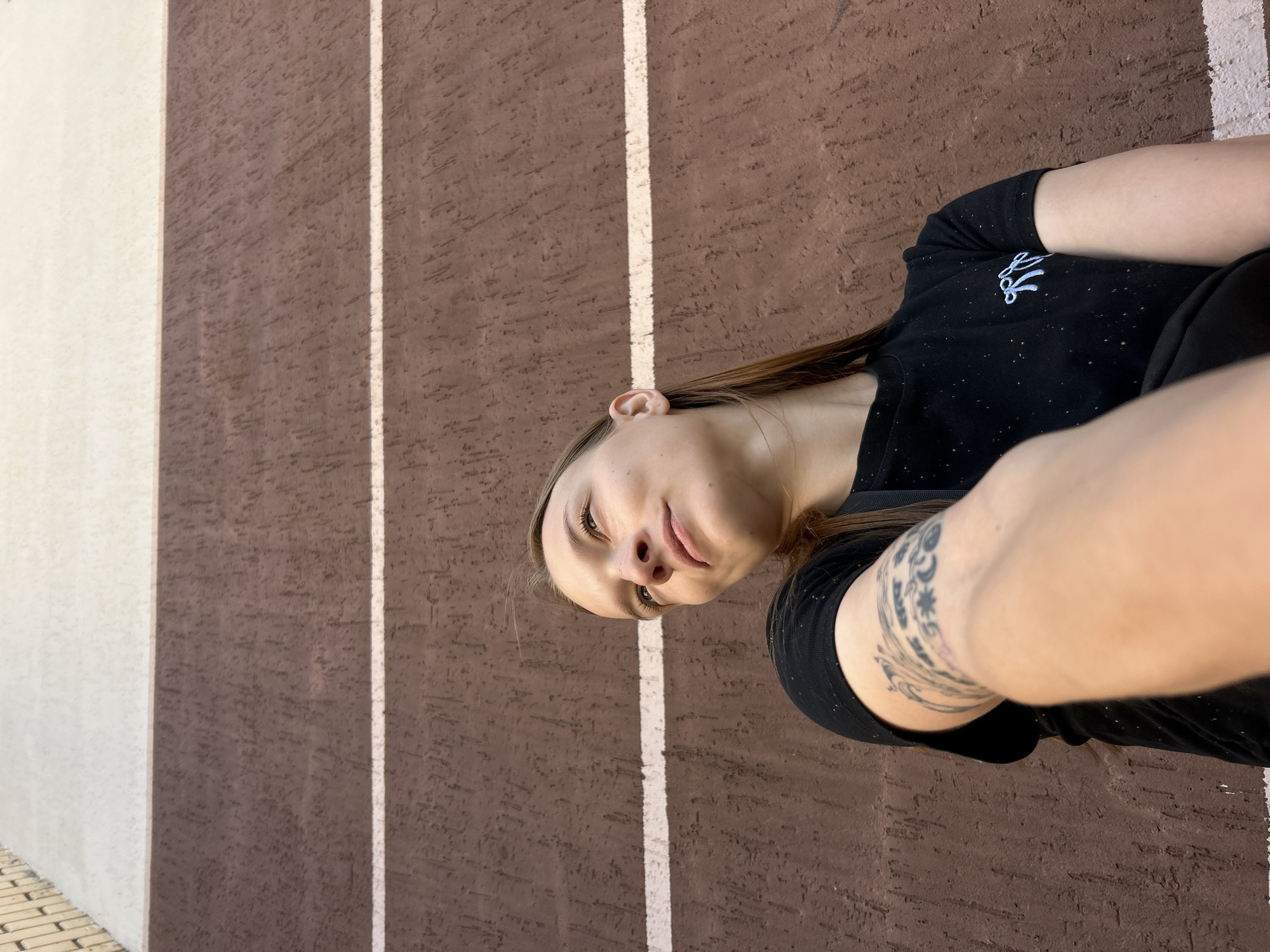 A young woman with long brown hair is shown from a low angle, looking upwards and to her left. She is wearing a black t-shirt with a small, light blue embroidered bow on the left side of the chest. She has tattoos on her left forearm. She is leaning against a textured brown wall with horizontal white stripes. The wall appears to be the exterior of a building in the city of Chaikovsky, Russia. The upper portion of the image shows a lighter colored section of the building, possibly concrete or stucco. The lighting suggests it is daytime.