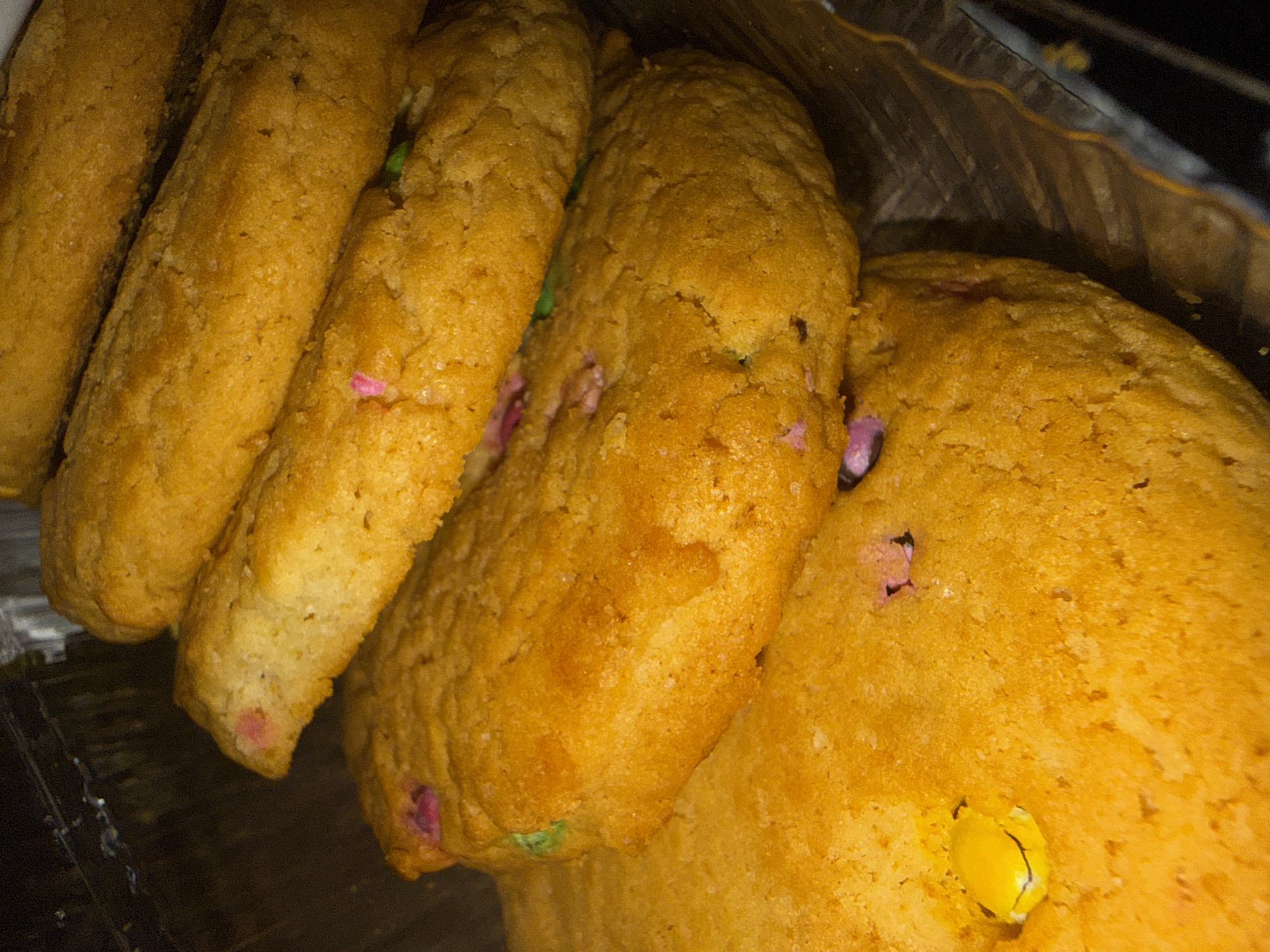 A close-up photograph depicts a collection of cookies, likely arranged inside a plastic container. The cookies appear to be a golden-brown color and contain what appear to be multicolored candies or sprinkles. The cookies vary in shape, with some being more elongated and others more round. The container shows some signs of being plastic. The scene does not contain any information related to Lupanovo, Russia. There are no people visible in the image.