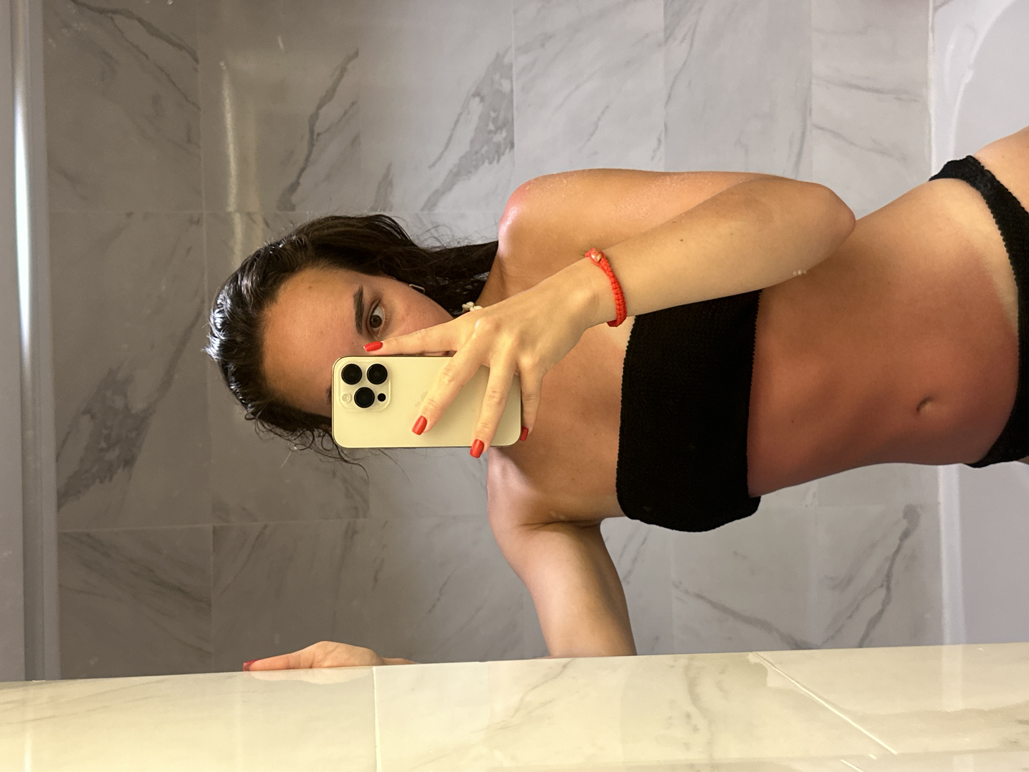A woman with wet dark hair is taking a selfie in a bathroom with marble-patterned walls. She is wearing a black bandeau top and a black bikini bottom. The woman has a red bracelet on her left wrist and red nail polish. Her face is partially obscured by a gold-colored smartphone. The scene appears to be a bathroom with a white countertop. The location is Pattaya City, Thailand.