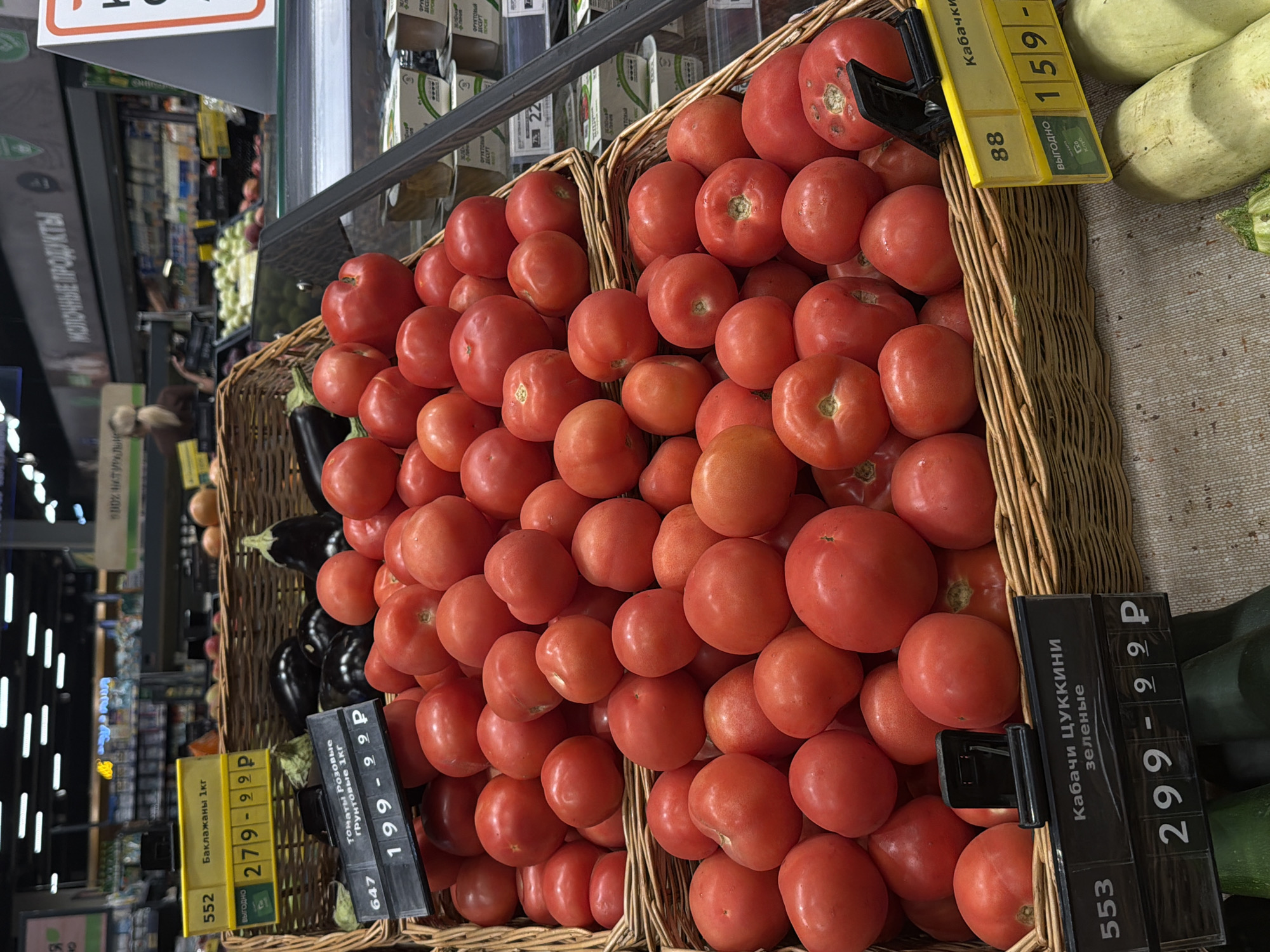 The image shows a close-up view of a produce display in a supermarket. In the foreground, there are two woven baskets filled with ripe red tomatoes. One basket is placed on top of another. A black price tag holder with a price of 299 ₽ for "Кабачи цуккини зеленые" is visible in the lower left, and a yellow price tag holder with a price of 159 ₽ for "Кабачки" is visible in the lower right. Behind the tomatoes, in another woven basket, are several dark purple eggplants. In the background, shelves stocked with various food items and products can be seen, along with some blurred figures of shoppers and store employees. The store appears to be a large supermarket chain. The location is a supermarket in the city of Perm, Russia.