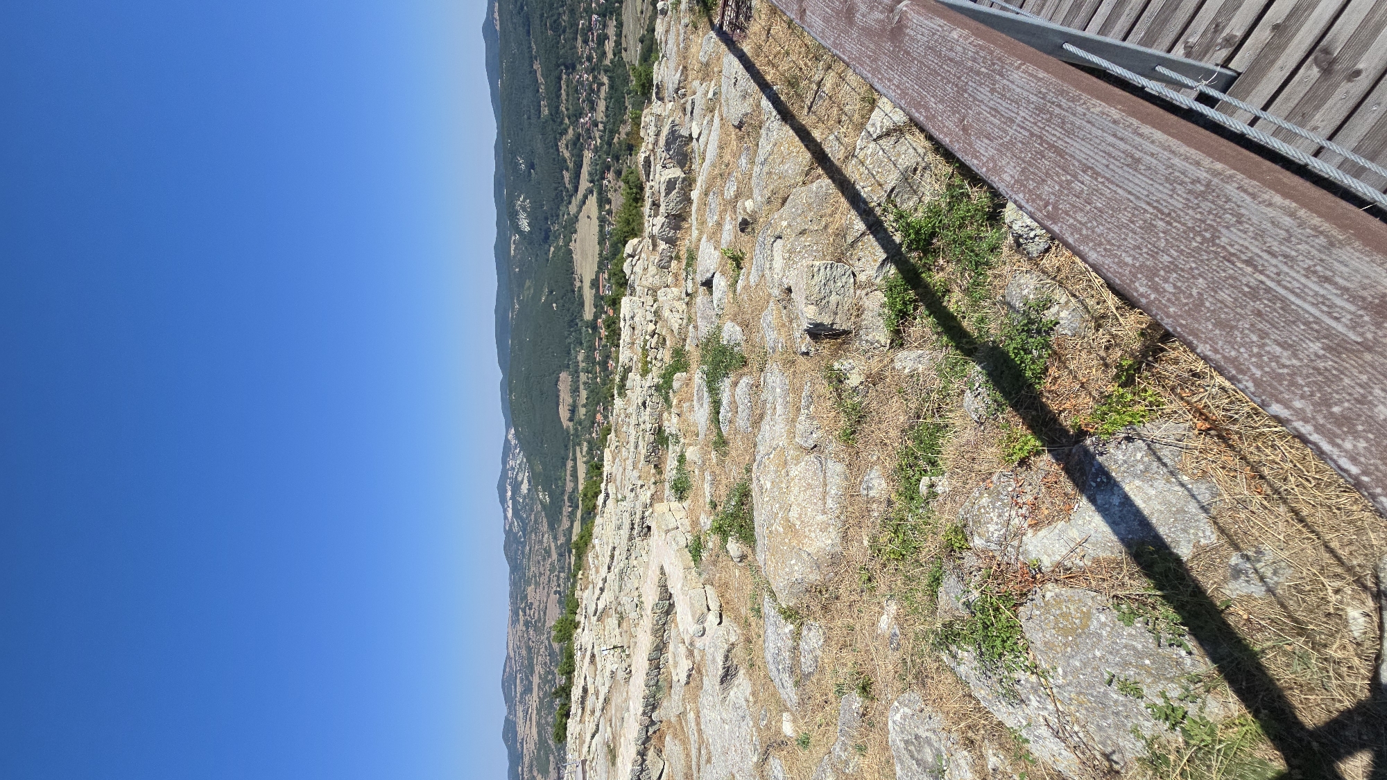 The image displays a panoramic view of ancient ruins and the surrounding landscape under a clear blue sky. In the foreground, a wooden railing and walkway suggest a viewing platform. The ground is covered with weathered stone structures, indicative of an archaeological site, interspersed with dry grass and sparse green vegetation. The middle ground reveals a valley with rolling hills covered in dense green forests and dotted with scattered houses, forming a village. The distant background is characterized by more hills and mountains. The scene captures a sunny day in the historical complex of Gorna Krepost, Bulgaria, highlighting the preserved remnants of a past civilization within a natural, mountainous setting.
