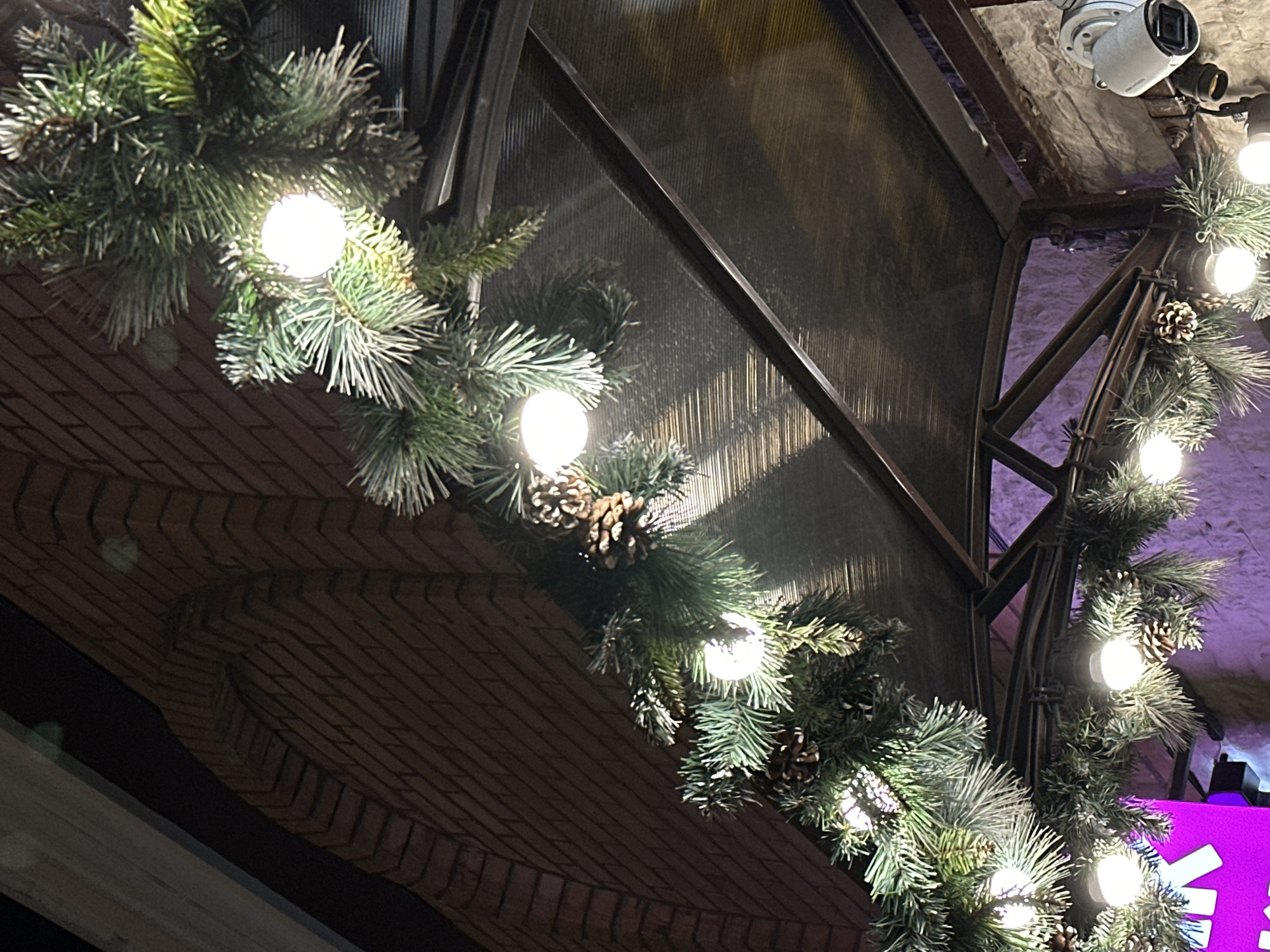 A low-angle shot shows a building adorned with lighted pine garlands, likely for a festive occasion. The building features brickwork and a metal support structure, with a security camera visible near the top right. The garlands are interspersed with pine cones and illuminated by spherical lights. In the background, a blurred purple hue suggests additional lighting or signage. The location could be Moscow, Russia, given the urban architectural style and the possible festive context.