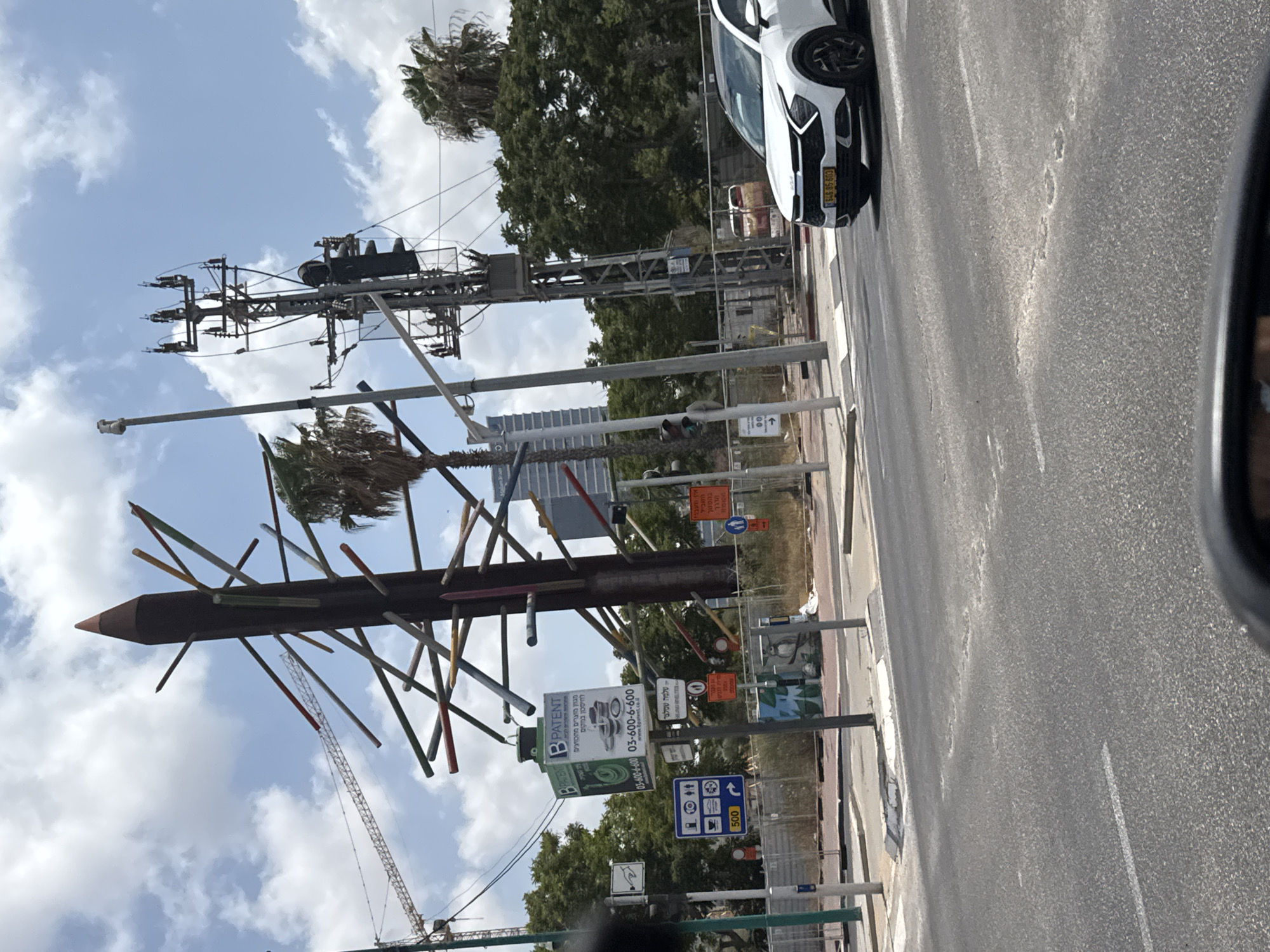 The image captures a street scene in Petah Tikva, Israel, on a bright, cloudy day. Dominating the background is a large, dark, pencil-shaped sculpture adorned with numerous protruding sticks of various colors. Adjacent to this sculpture, a tall electrical pole with transformers and what appears to be a traffic light stands prominently. In the middle ground, a white SUV with a yellow license plate is visible on the road, indicating it is in motion or stopped at an intersection. The foreground consists of an asphalt road with lane markings and a pedestrian crossing. Various signs are scattered throughout the scene, including a blue sign with icons for a fuel pump, a disabled person, and a toilet, as well as a sign with the number "500." The environment is urban, with trees and buildings in the background, and construction fencing along one side of the road. The overall impression is a typical urban streetscape with a notable art installation.