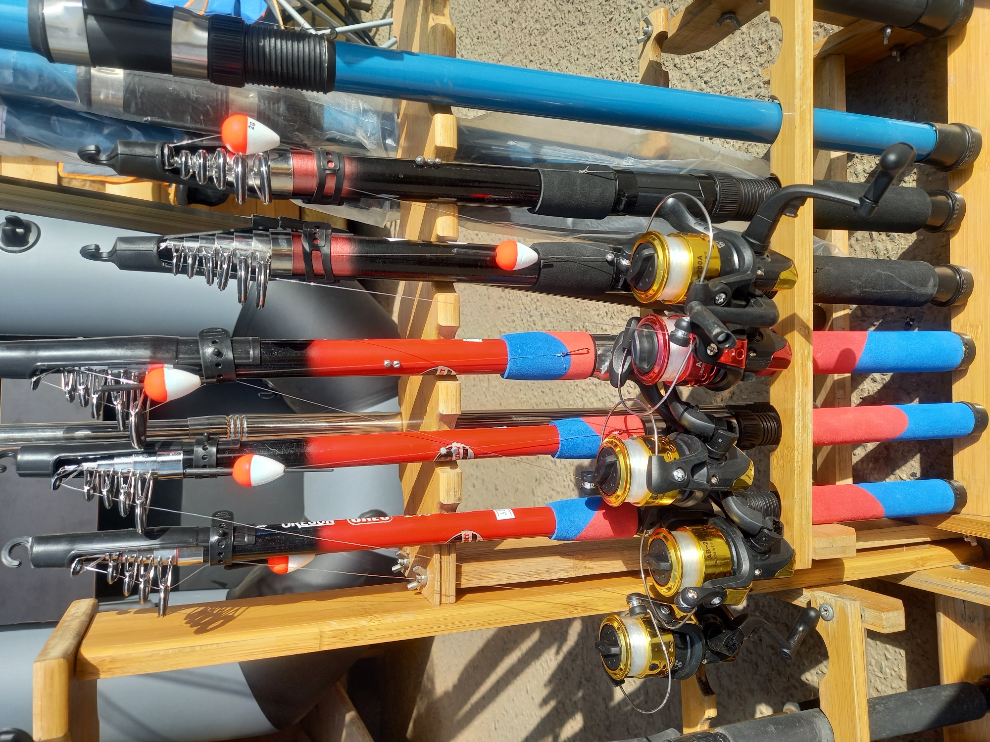 A wooden rack holds several fishing rods with reels. The rods are various colors, including blue, black, red, and silver. Some have floats attached. The reels are a mix of black, gold, and red. The rack is positioned outdoors under sunlight.